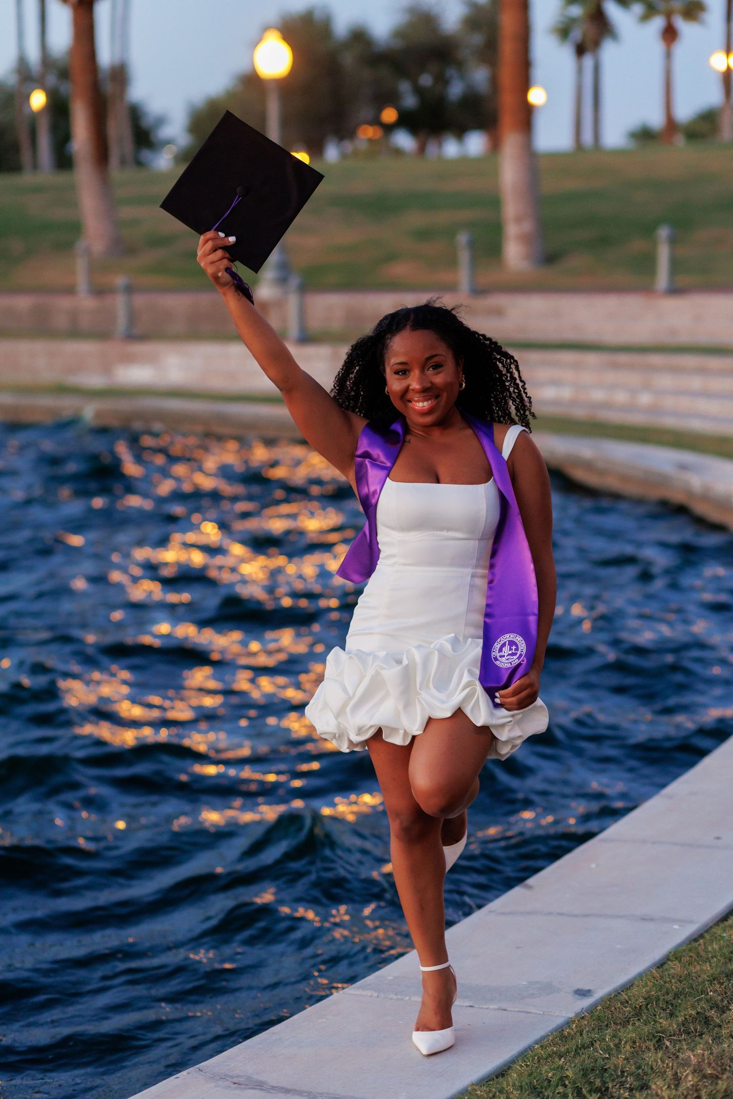 Joyful graduate in white dress raises black cap by reflective water at dusk with palm trees.