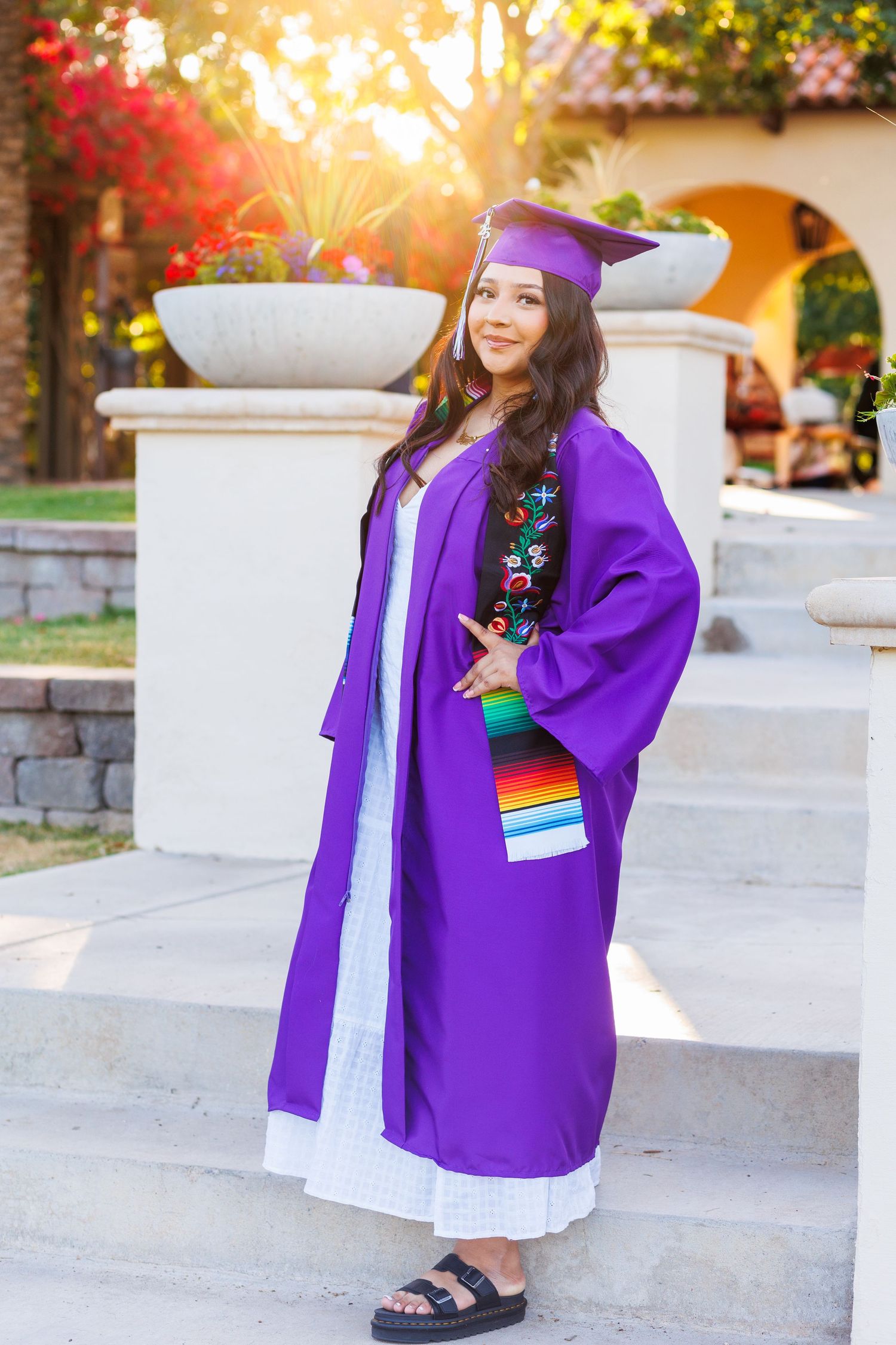 Young woman in purple graduation gown and cap posing on steps in golden sunlight.