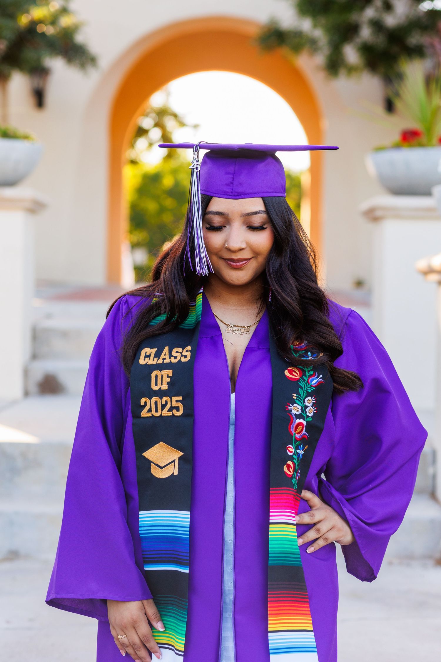 Graduate in purple cap and gown with Class of 2025 stole looking down outdoors.