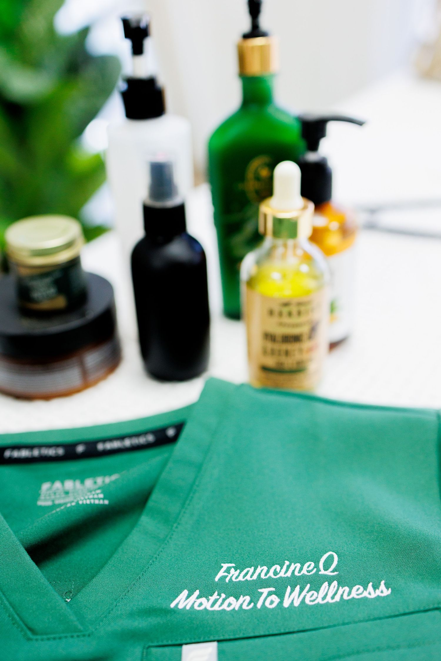 Green Fabletics scrubs top embroidered with Francine Q Motion To Wellness logo displayed near wellness products.