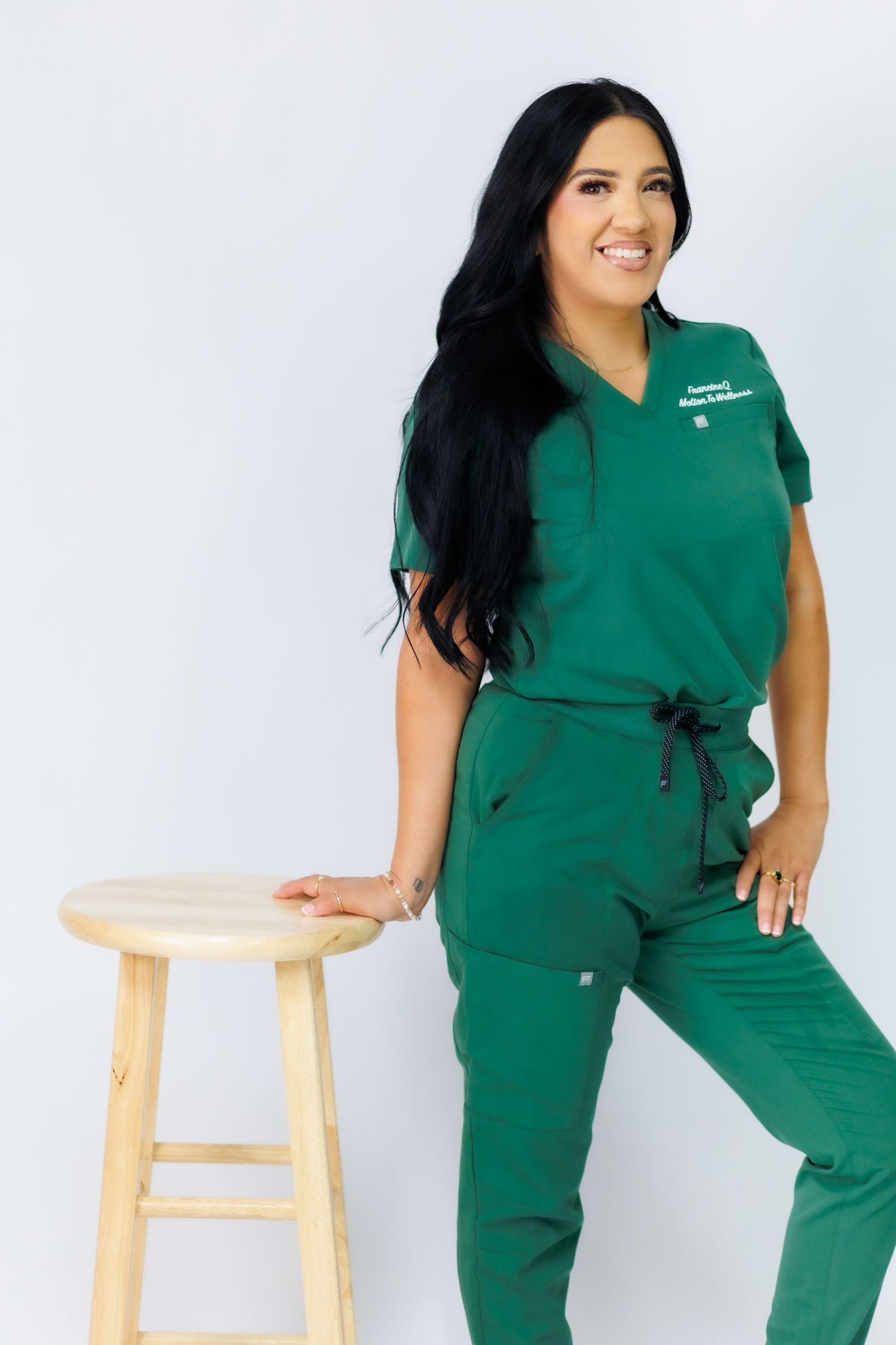 Woman in green Motion To Wellness scrubs uniform posing confidently beside a wooden stool against white background.