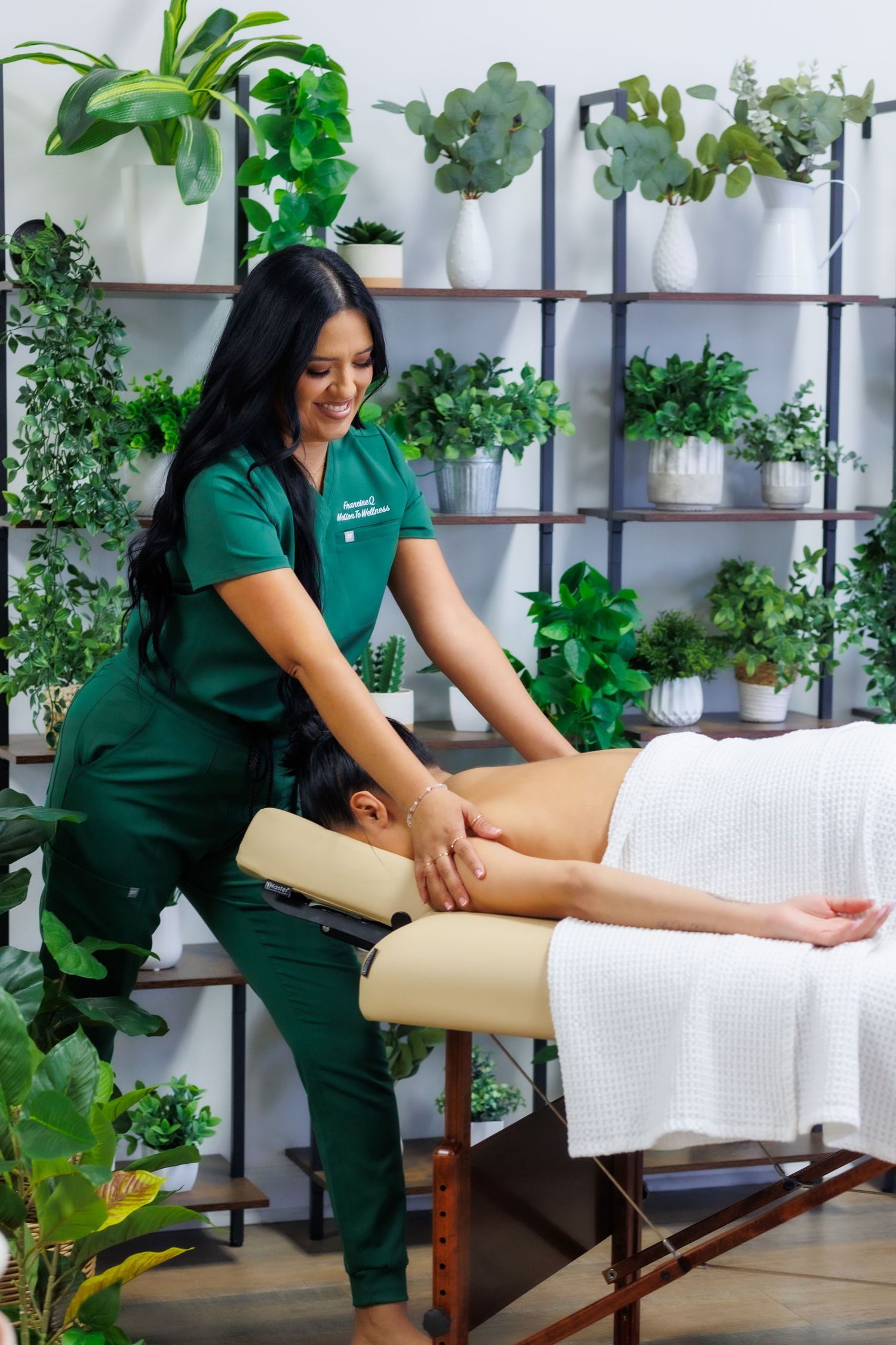 Licensed massage therapist in green scrubs performing back massage on client in plant-filled wellness studio.
