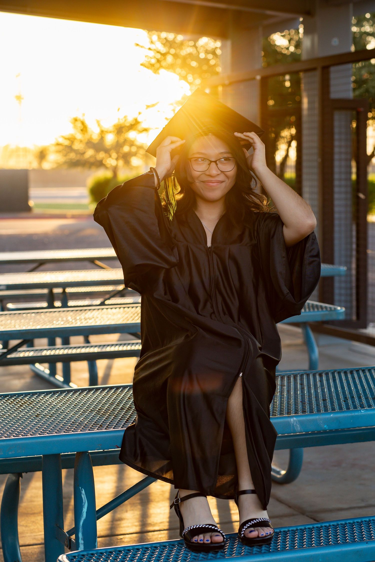 Young woman in black graduation cap and gown sitting outdoors at sunset, adjusting her cap and smiling.