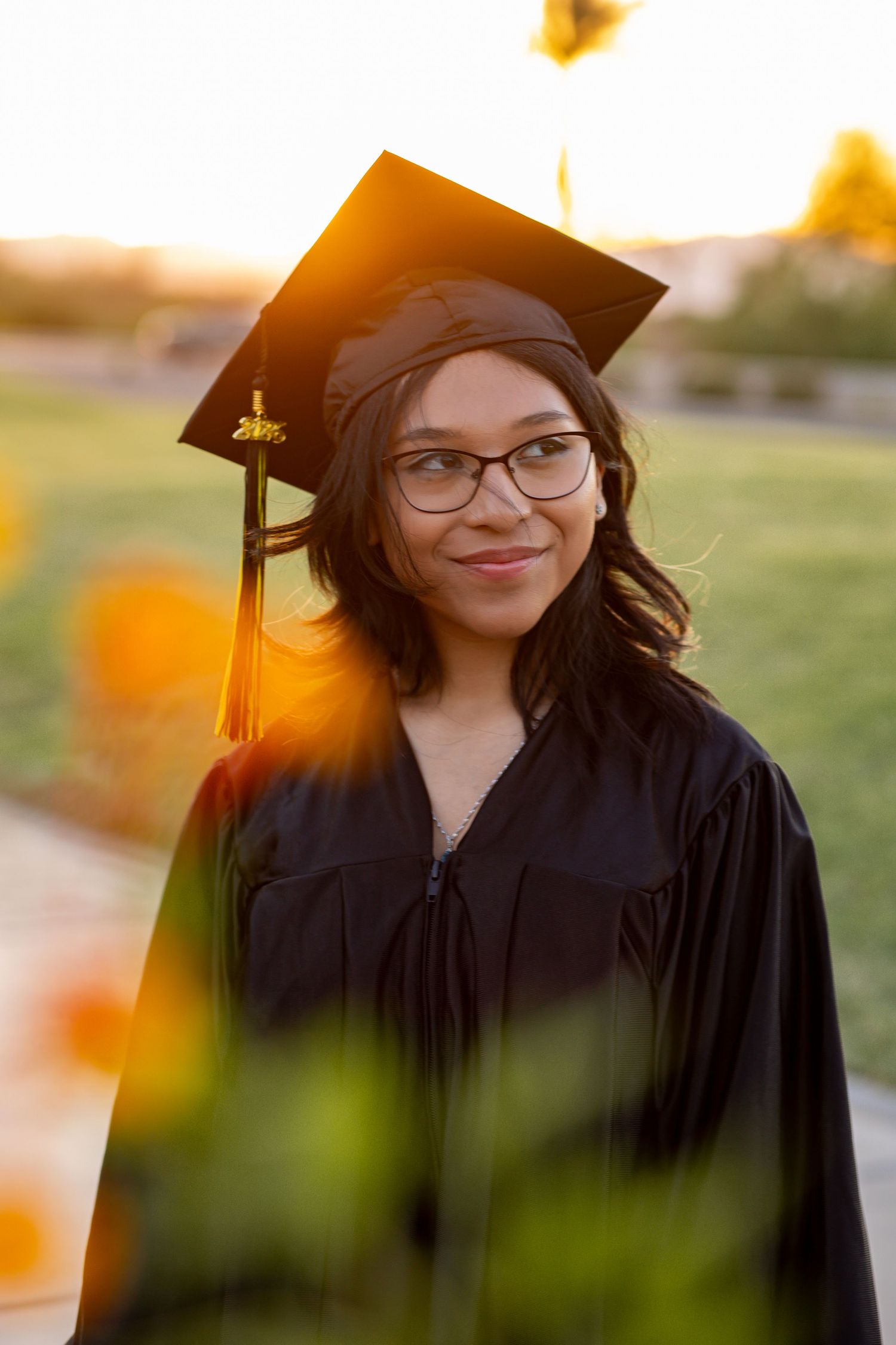 Young woman in black graduation cap and gown smiles outdoors at golden hour, wearing glasses.