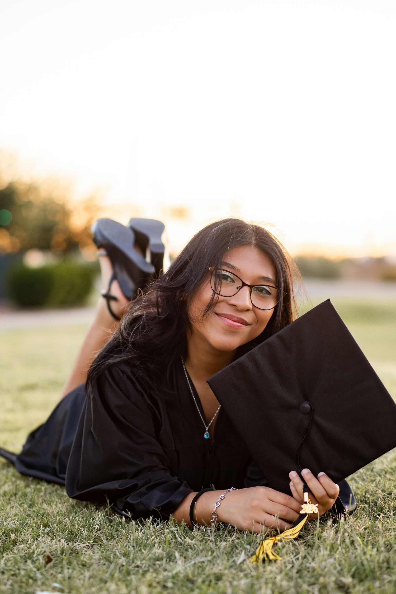 Young woman in black graduation gown and cap lying on grass, smiling at camera during golden hour.