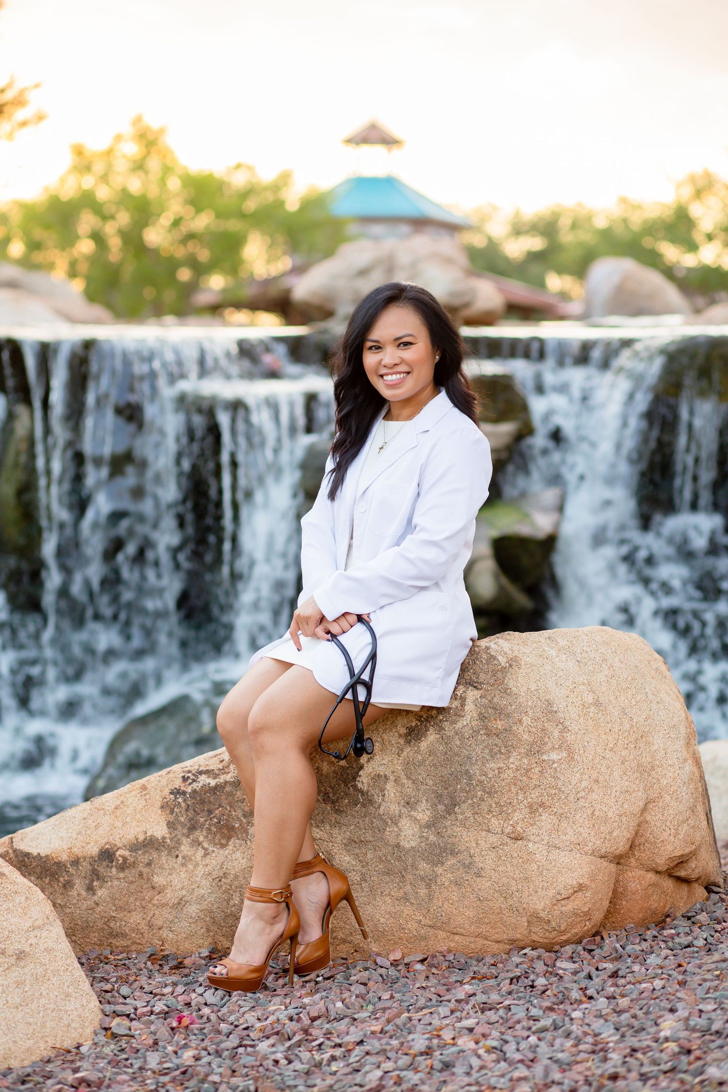 Female healthcare professional in white coat holding stethoscope sitting on rock near scenic waterfall outdoors.