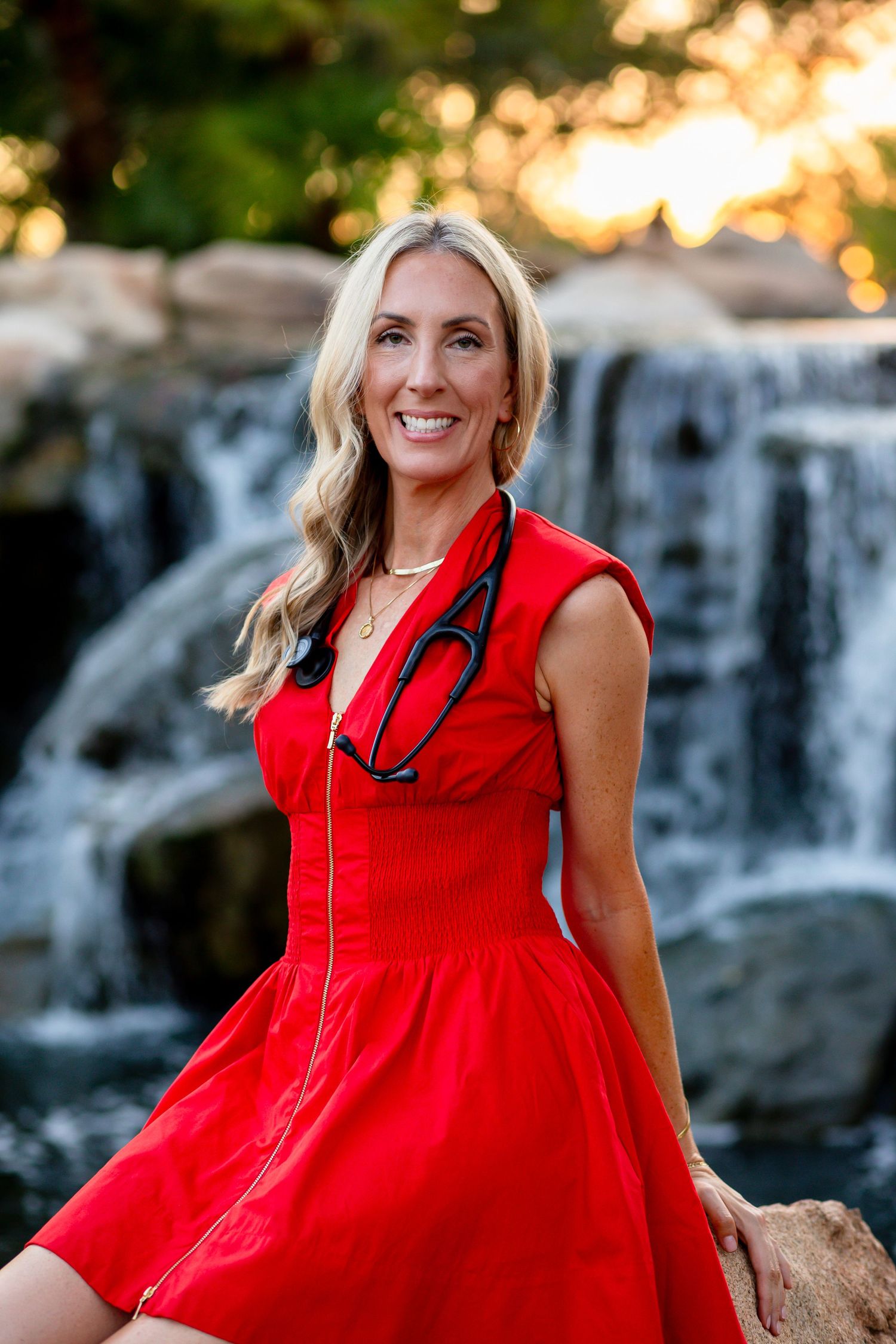 Smiling blonde woman in red dress wearing stethoscope posing near a waterfall at golden hour.