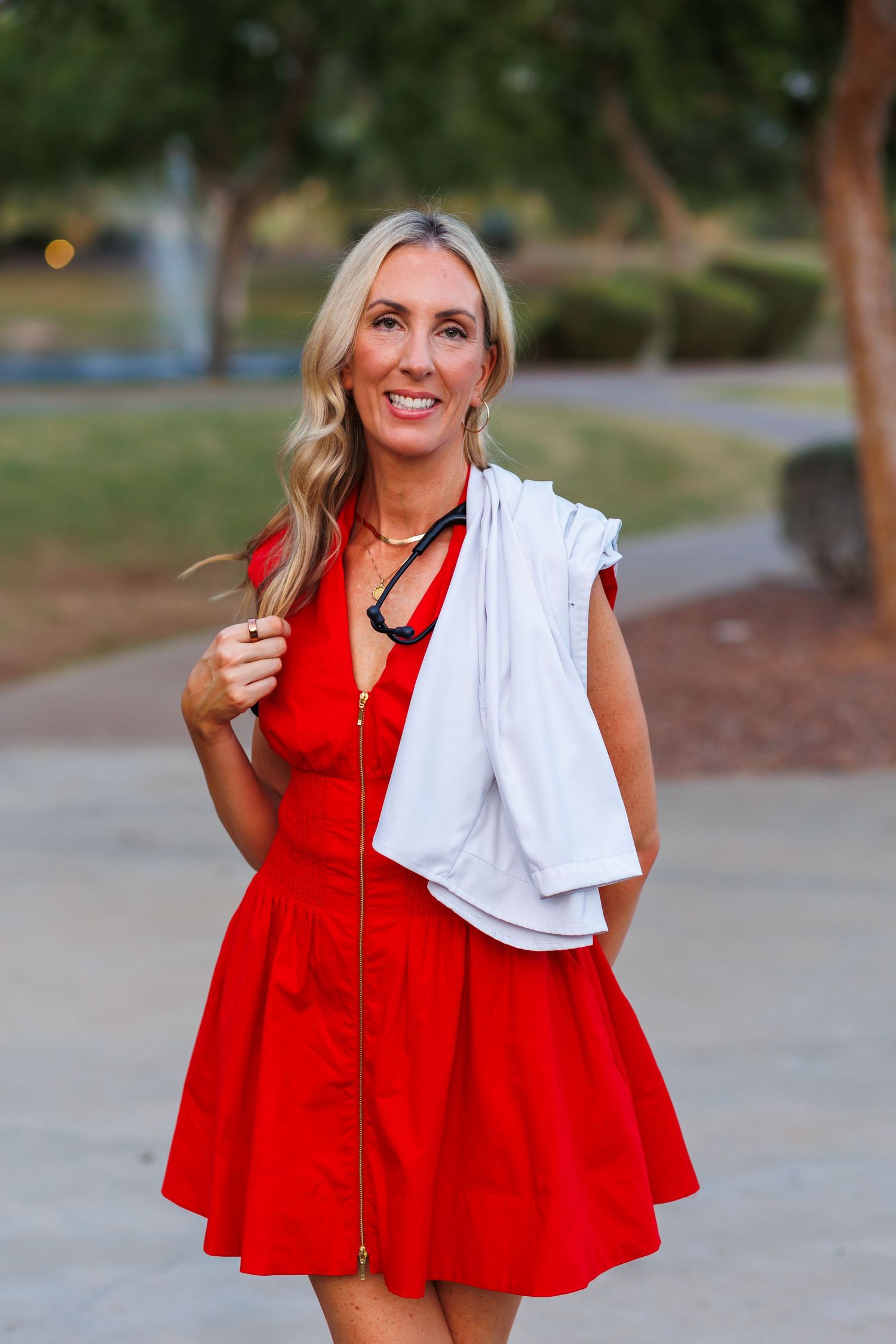 Cheerful blonde woman in red zip-up dress carrying white lab coat over shoulder in sunny park.
