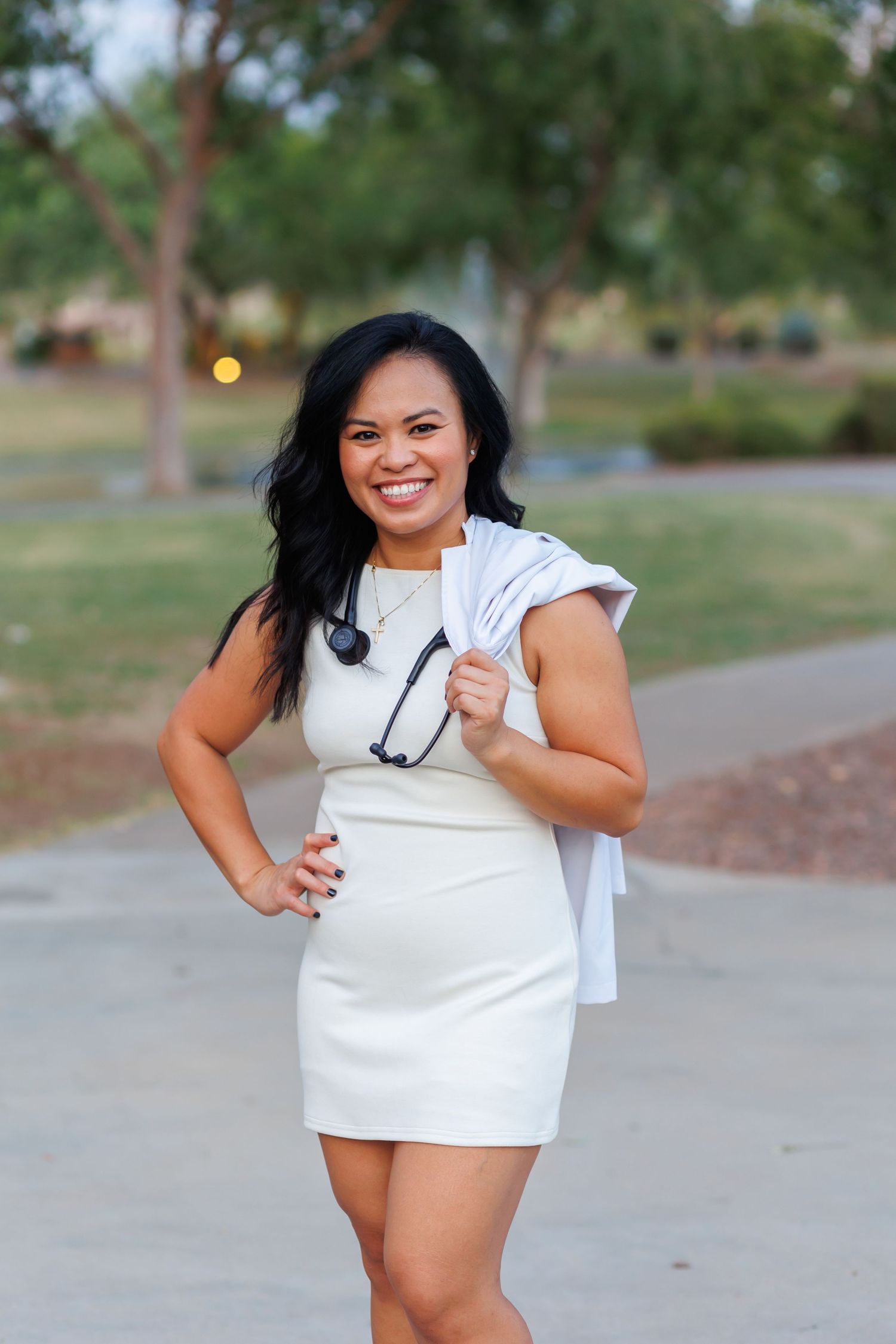 Smiling woman in white dress holding white coat and stethoscope outdoors in park setting.
