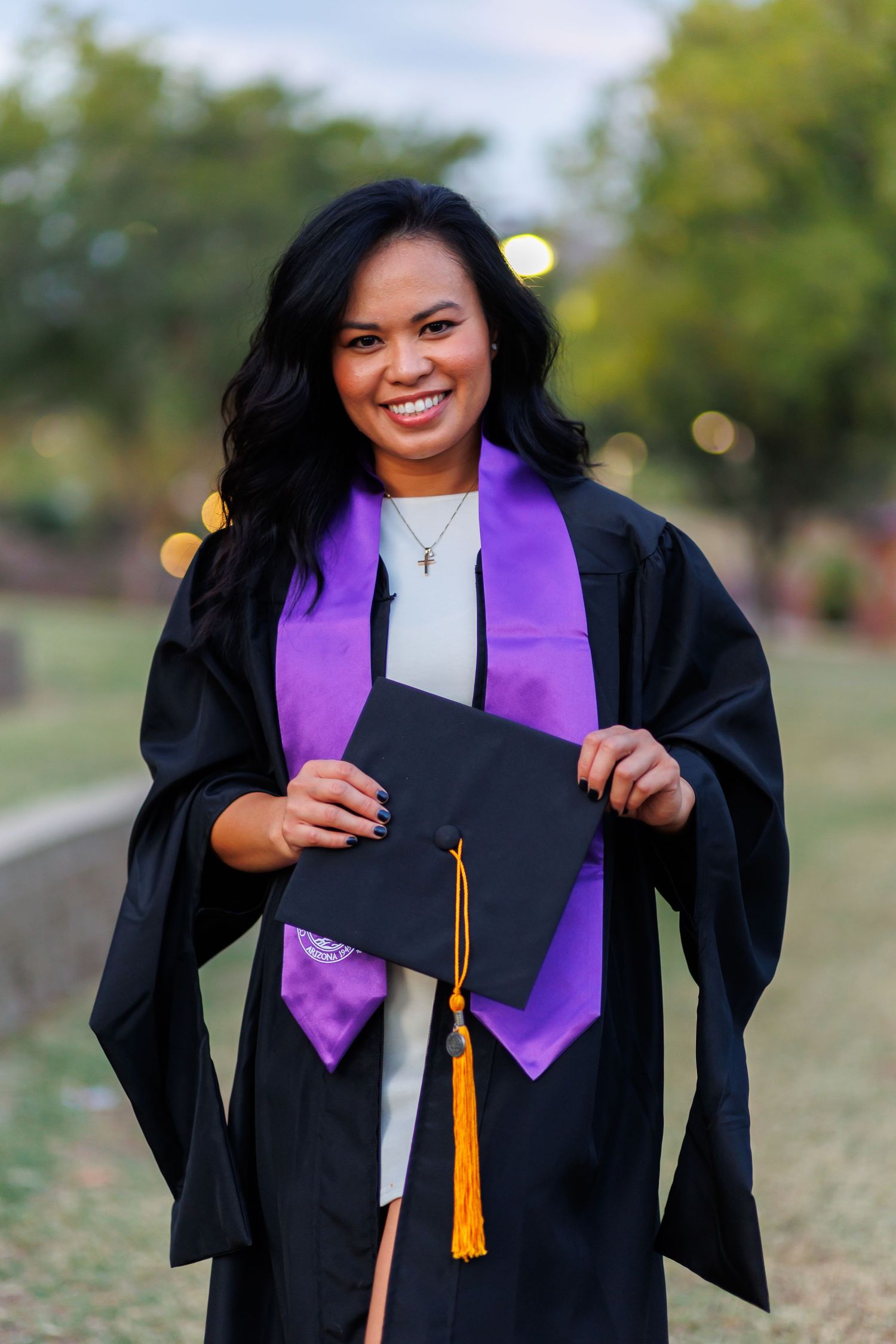 Smiling graduate in black gown with purple stole holding cap outdoors at graduation.