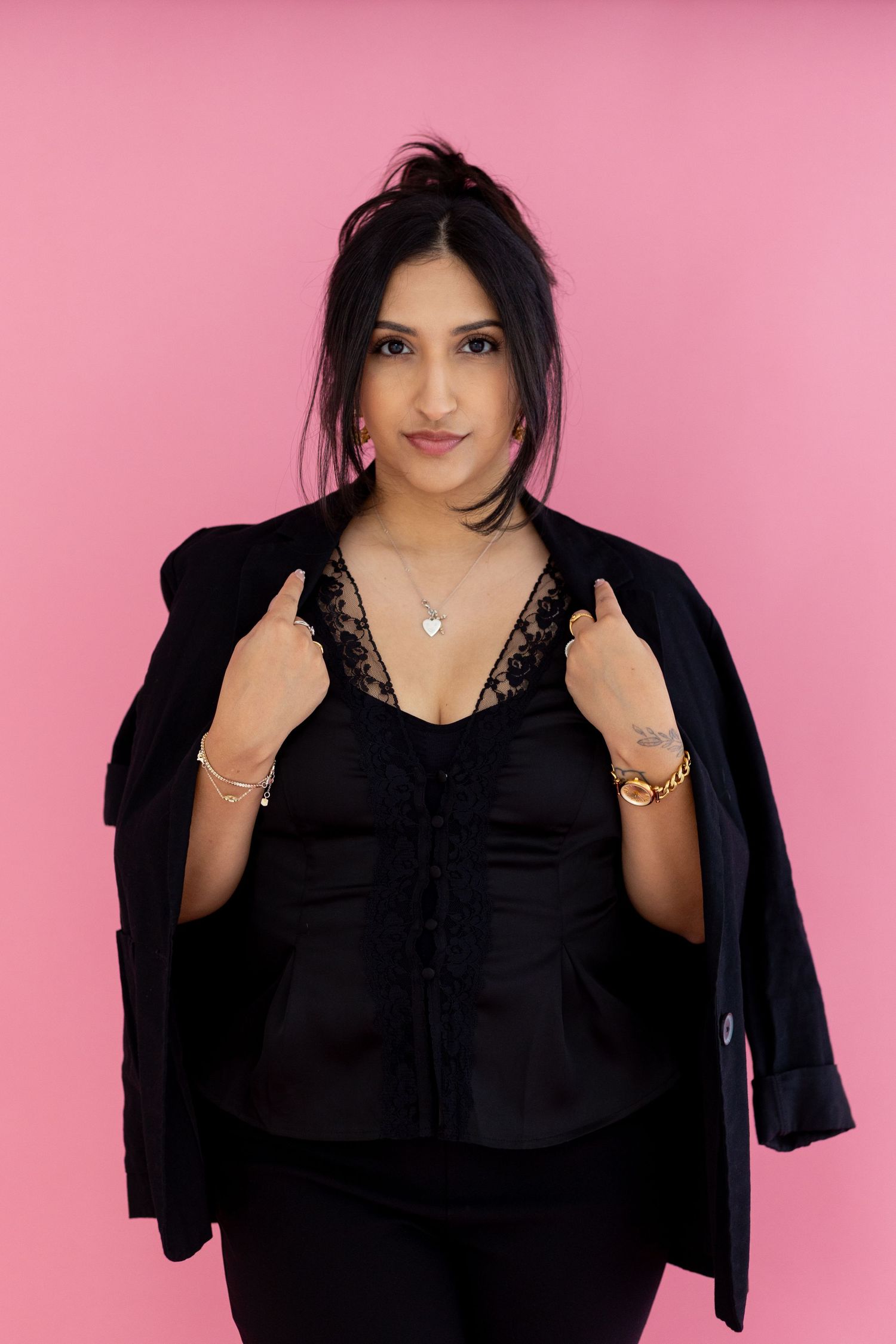 Woman in black lace top and blazer posing confidently against a pink background wearing gold jewelry.