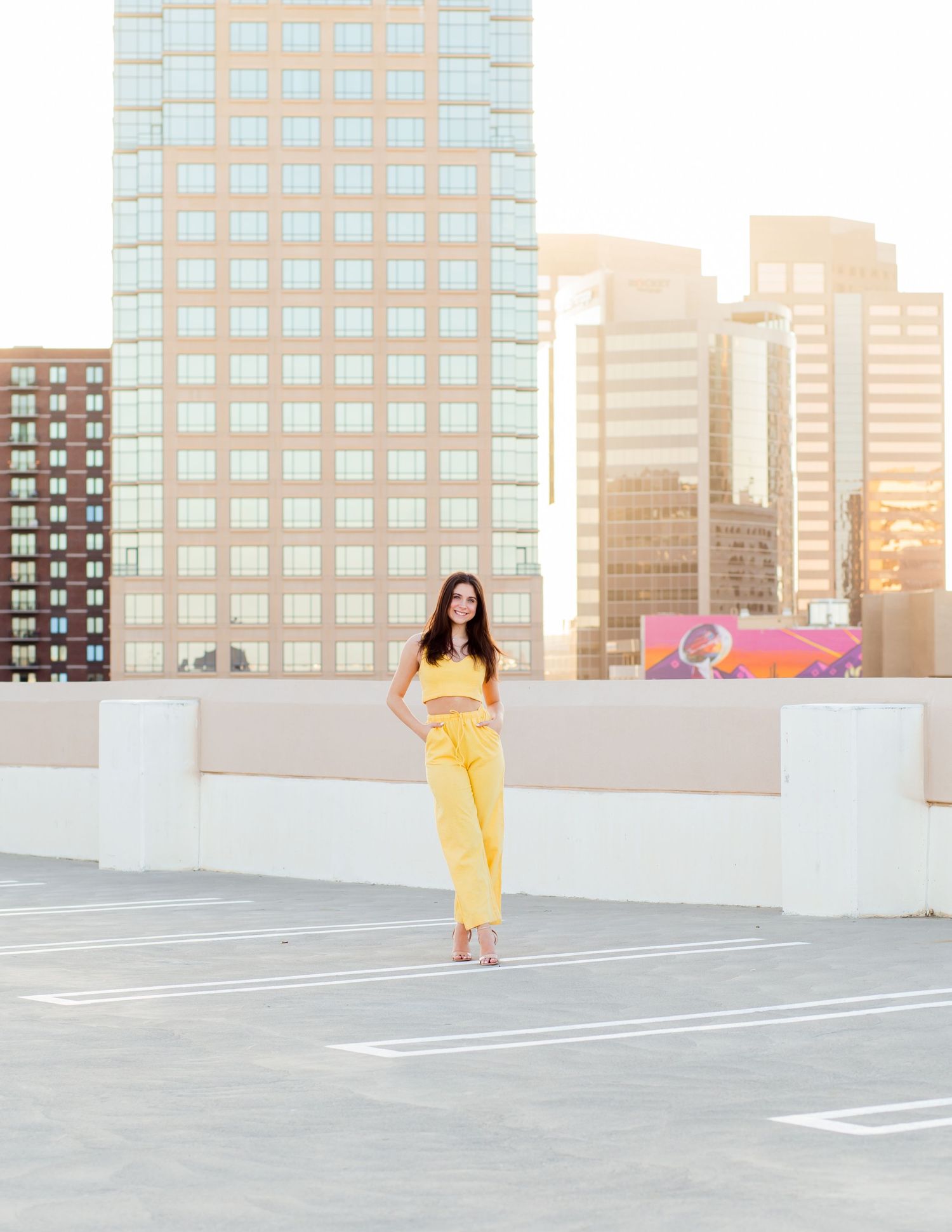 Young woman in yellow outfit stands on rooftop parking garage with city skyline at golden hour.