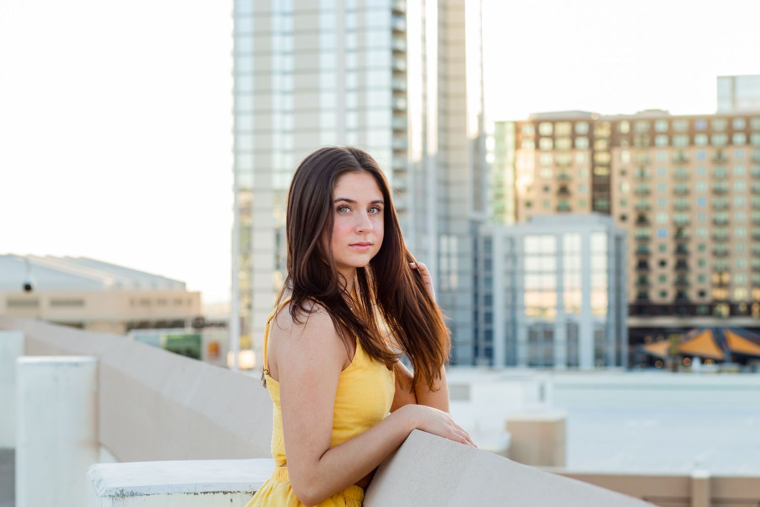 Brunette teen in yellow dress leans on rooftop wall with urban skyscrapers in background.