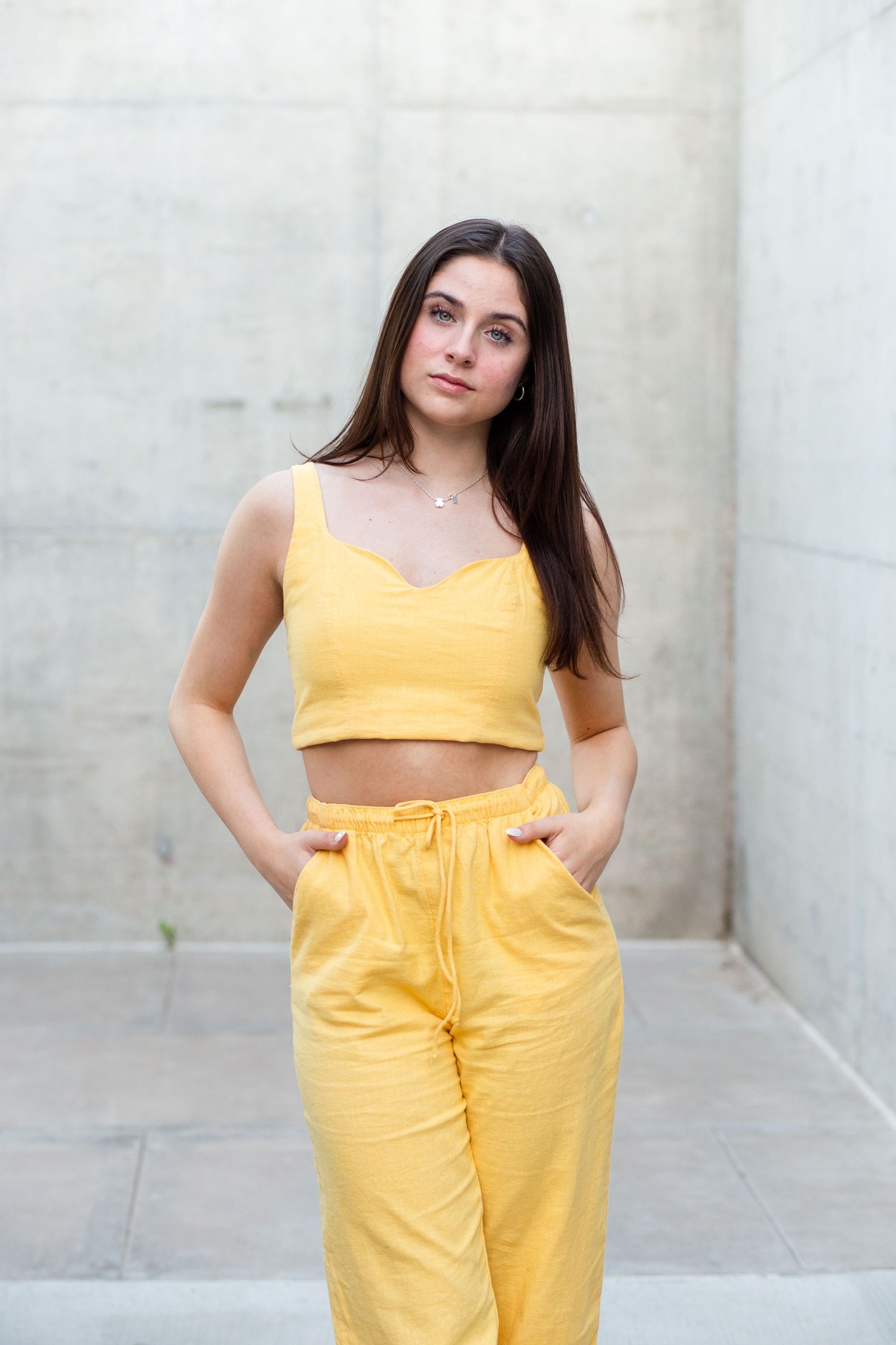 Young woman in yellow crop top and pants stands confidently against concrete wall outdoors.