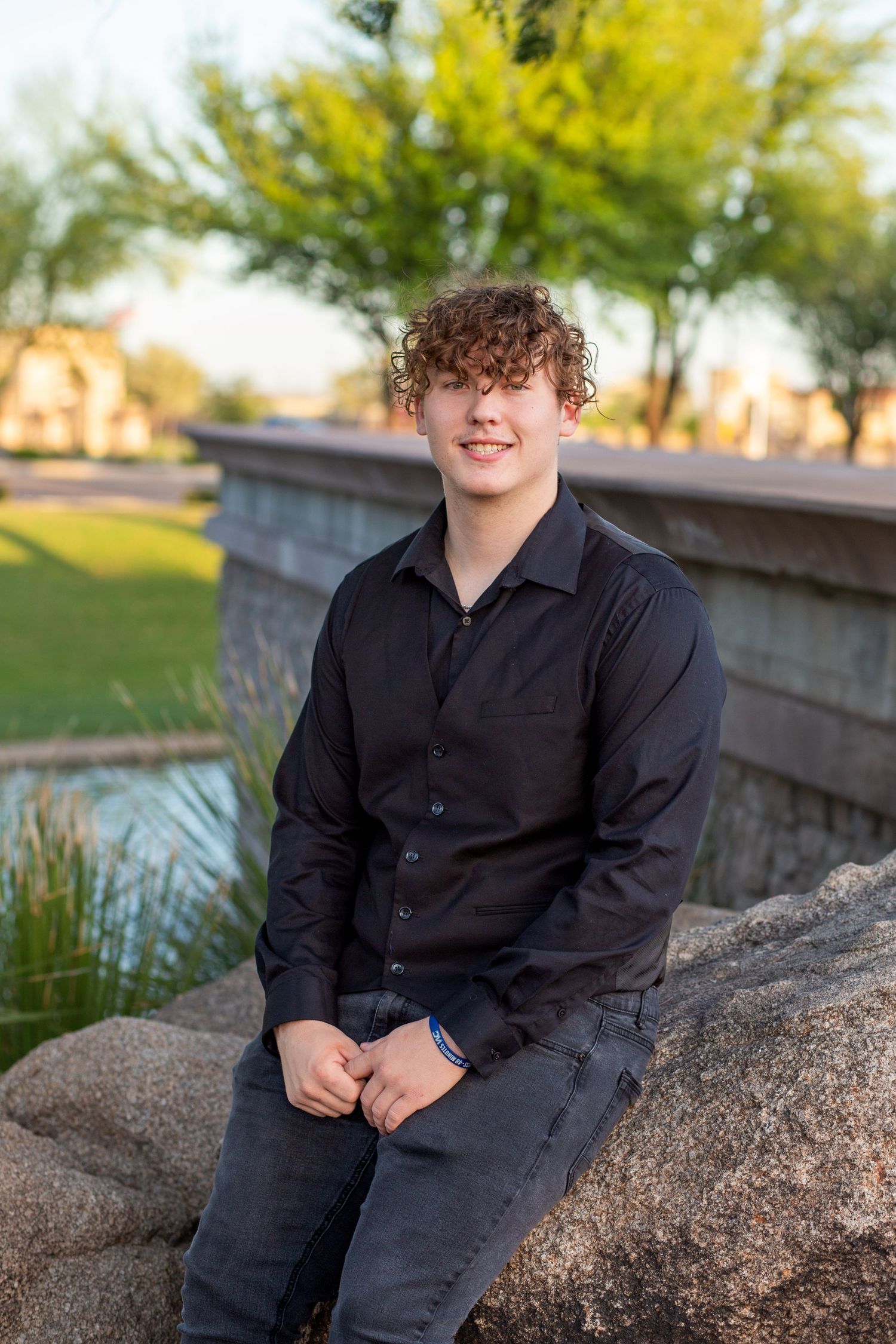 Young man with curly hair in black shirt sitting on rocks in park setting.