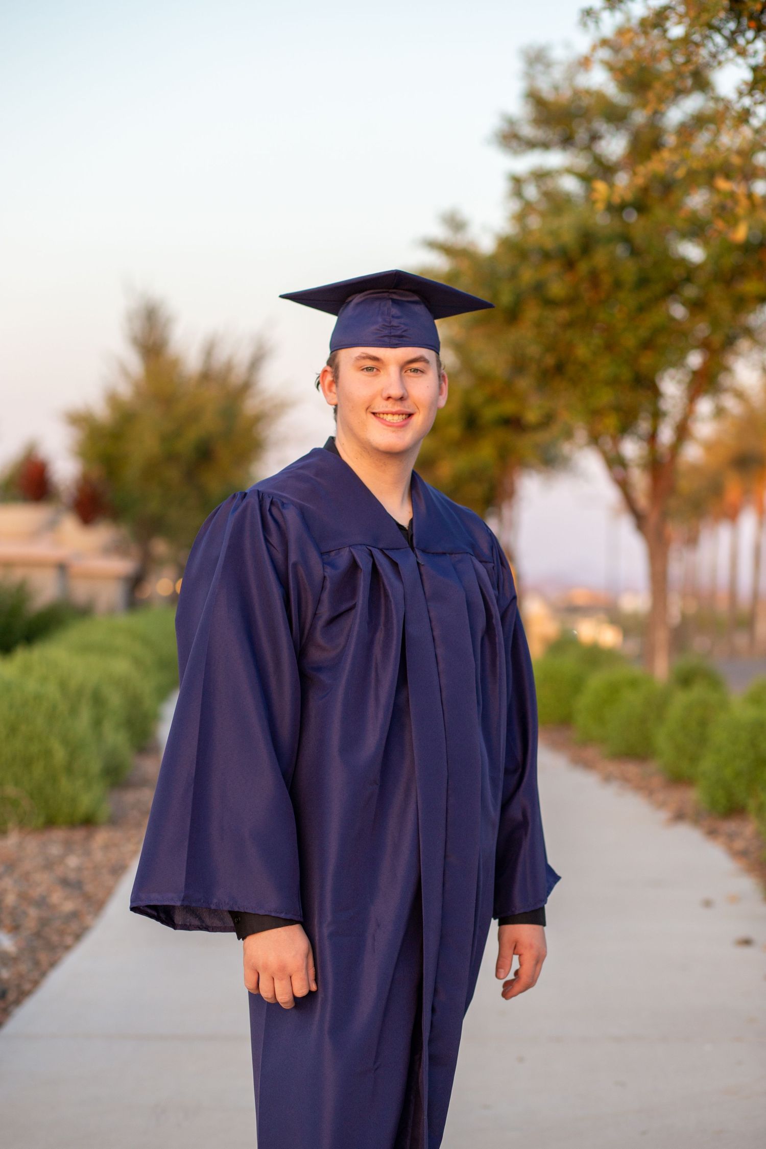 Young male graduate in navy blue cap and gown smiling on tree-lined pathway.