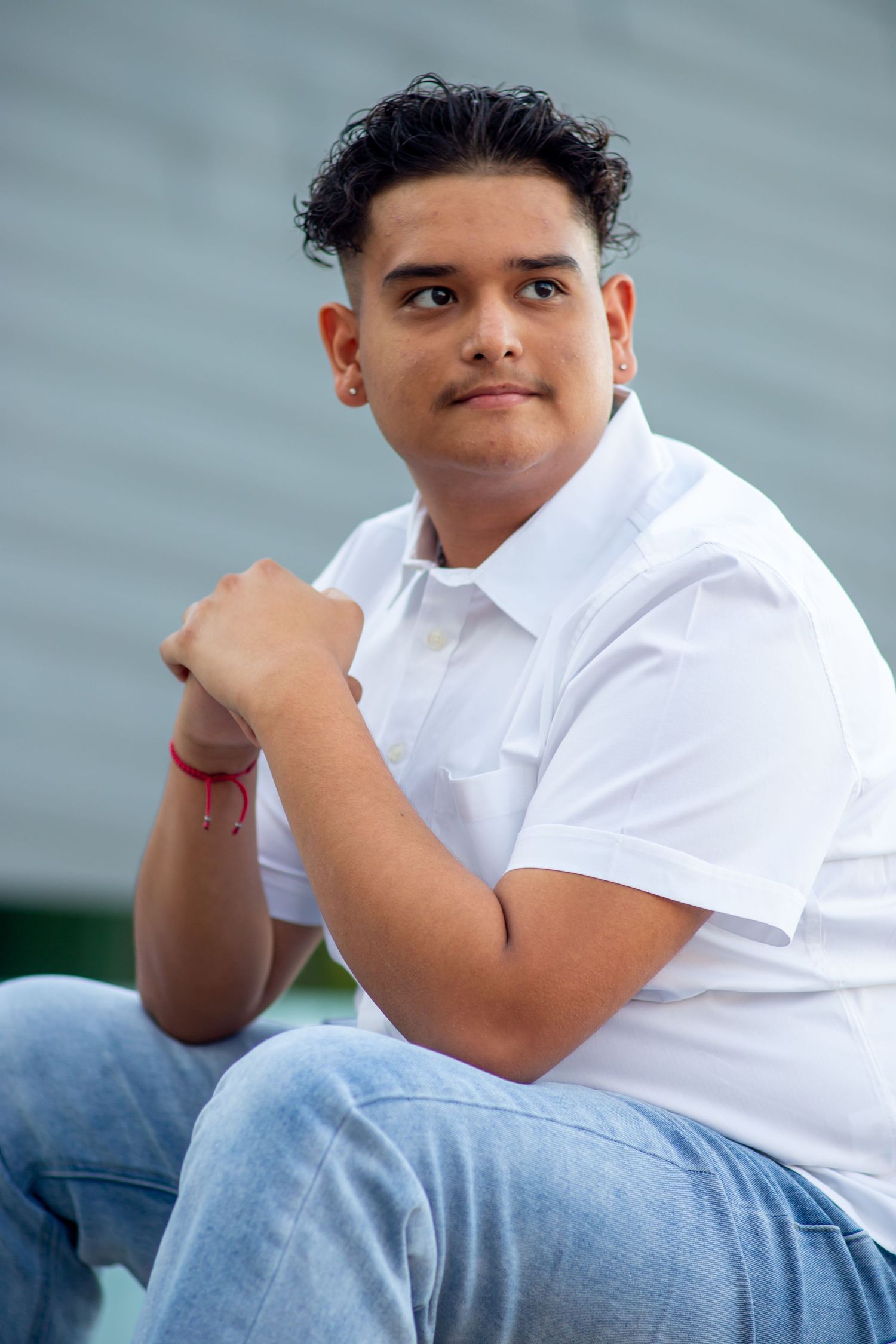 Young Hispanic man in white polo shirt sitting outdoors against gray wall background.