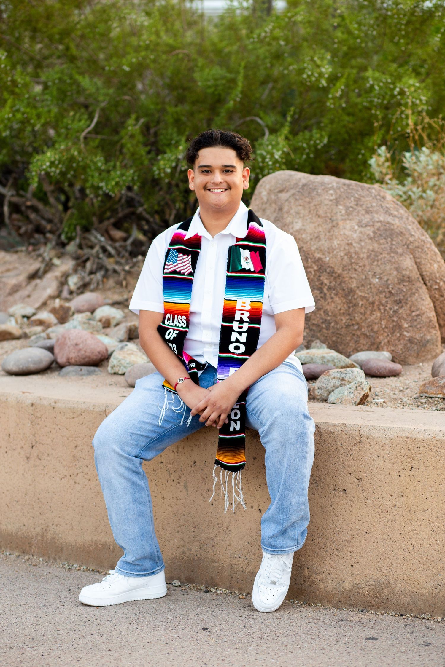 Young man in graduation sash with US and Mexican flags sits smiling on stone bench outdoors.