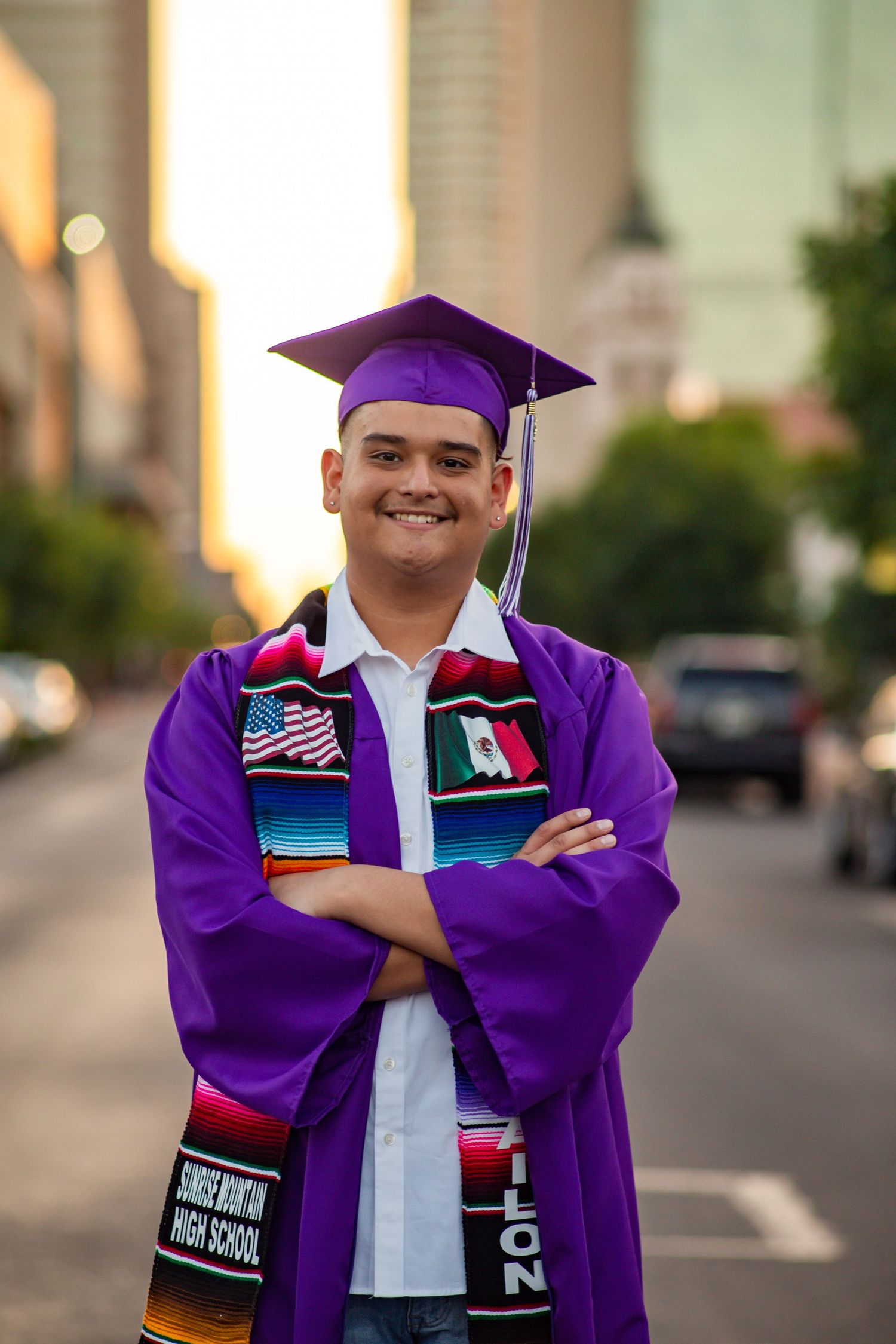Smiling graduate in purple cap and gown with colorful sash stands on city street at sunset.