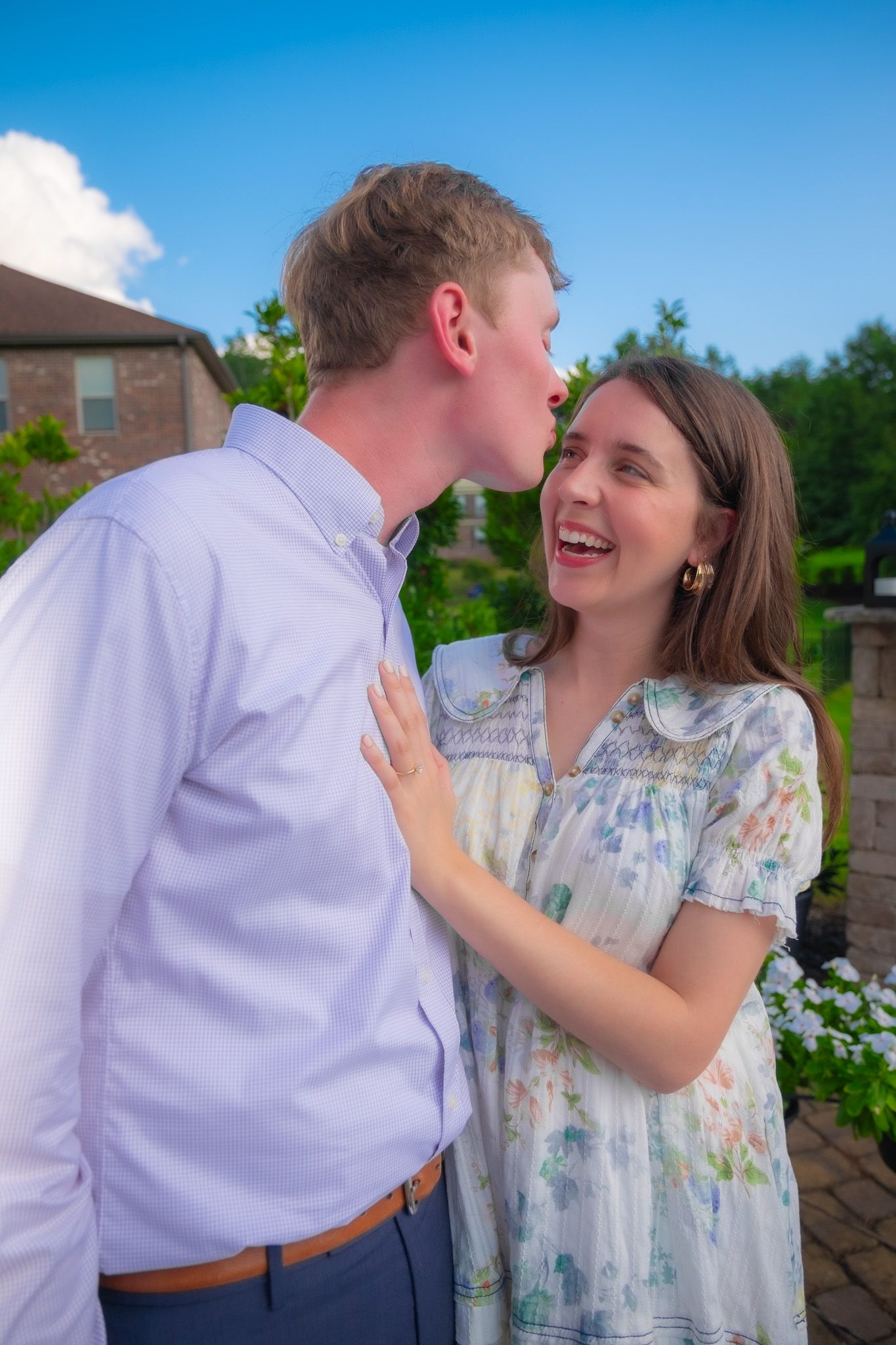 Couple shares joyful laughs and embraces during outdoor photo session in front of brick building with greenery.