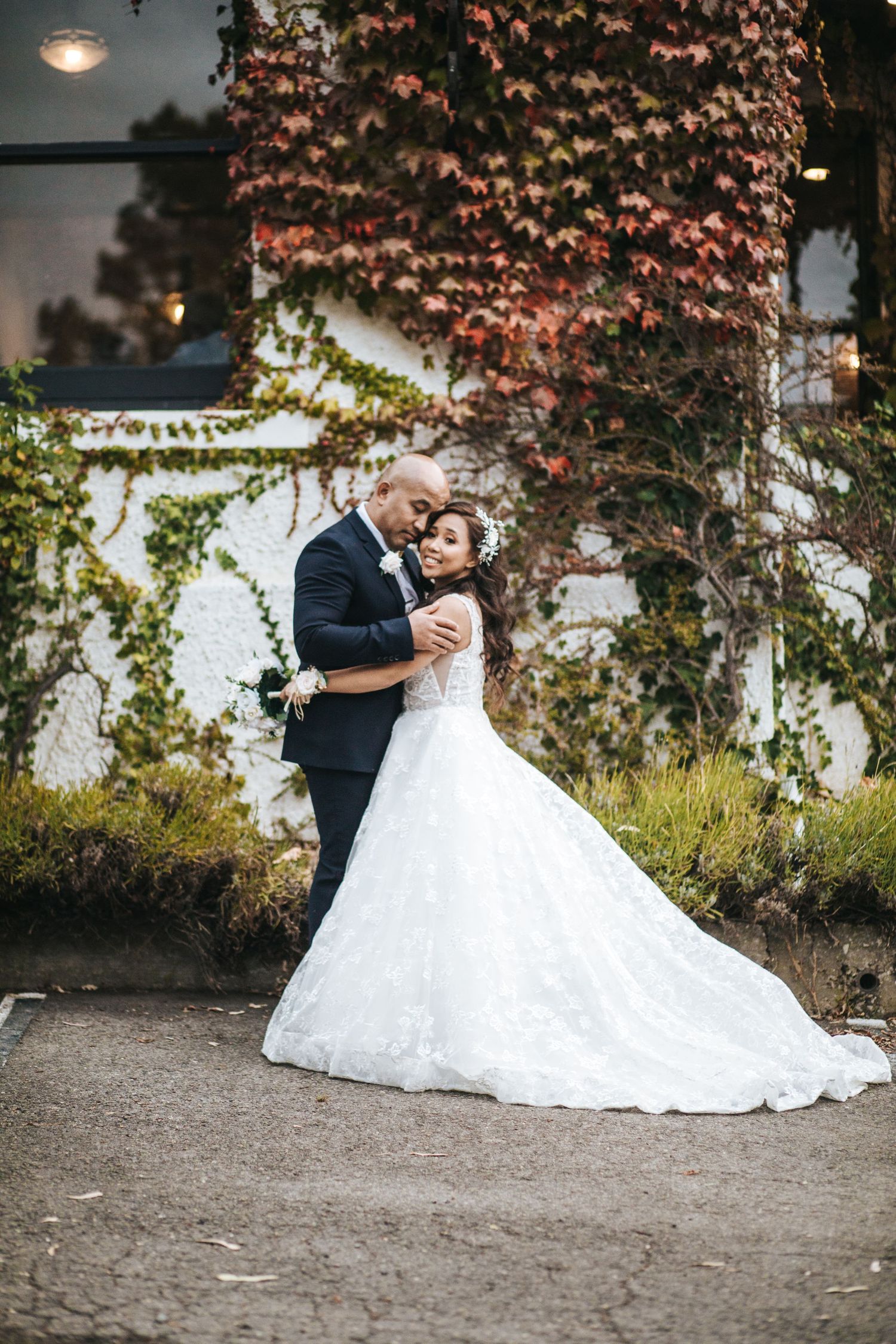 Elegant couple embraces in front of red autumn ivy-covered wall in classic wedding portrait.