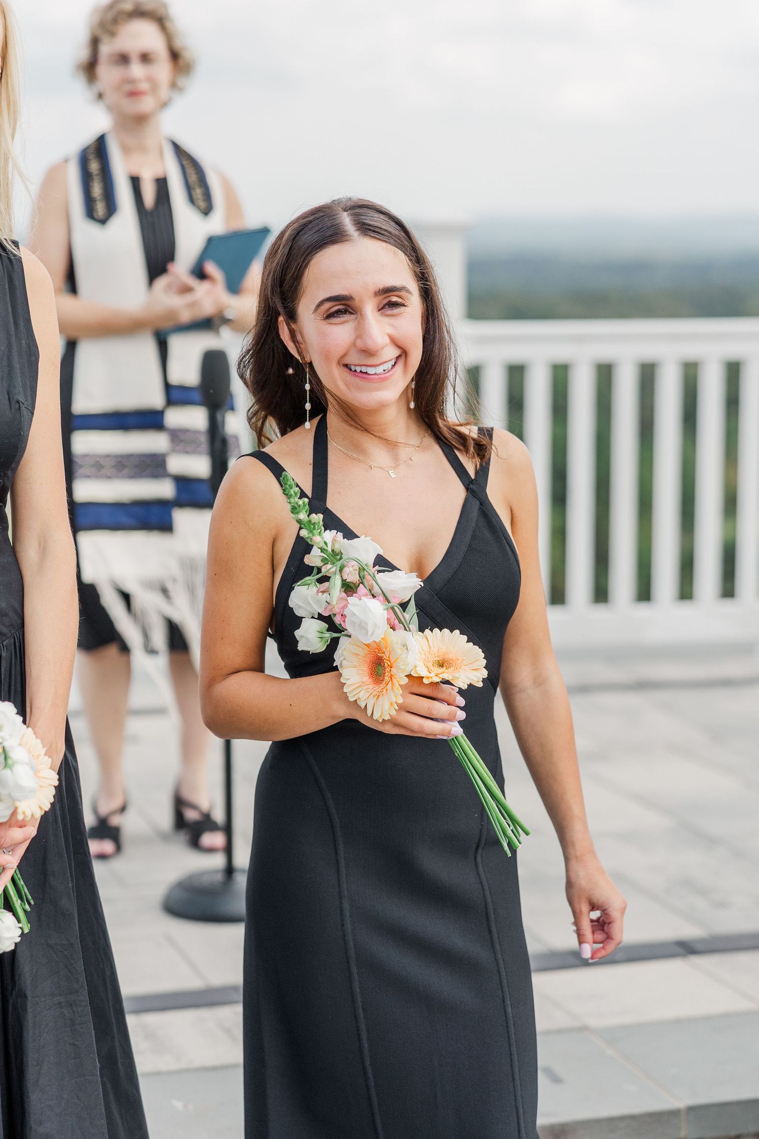 Wedding attendees in black dresses holding floral bouquets on stone terrace.