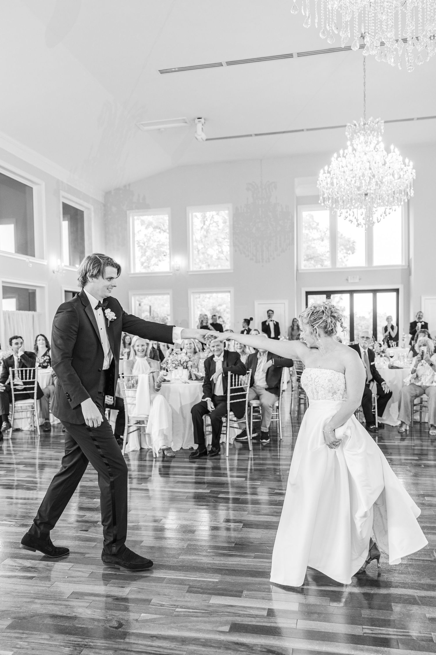 Black and white photo of a couple dancing in an elegant ballroom with crystal chandeliers.