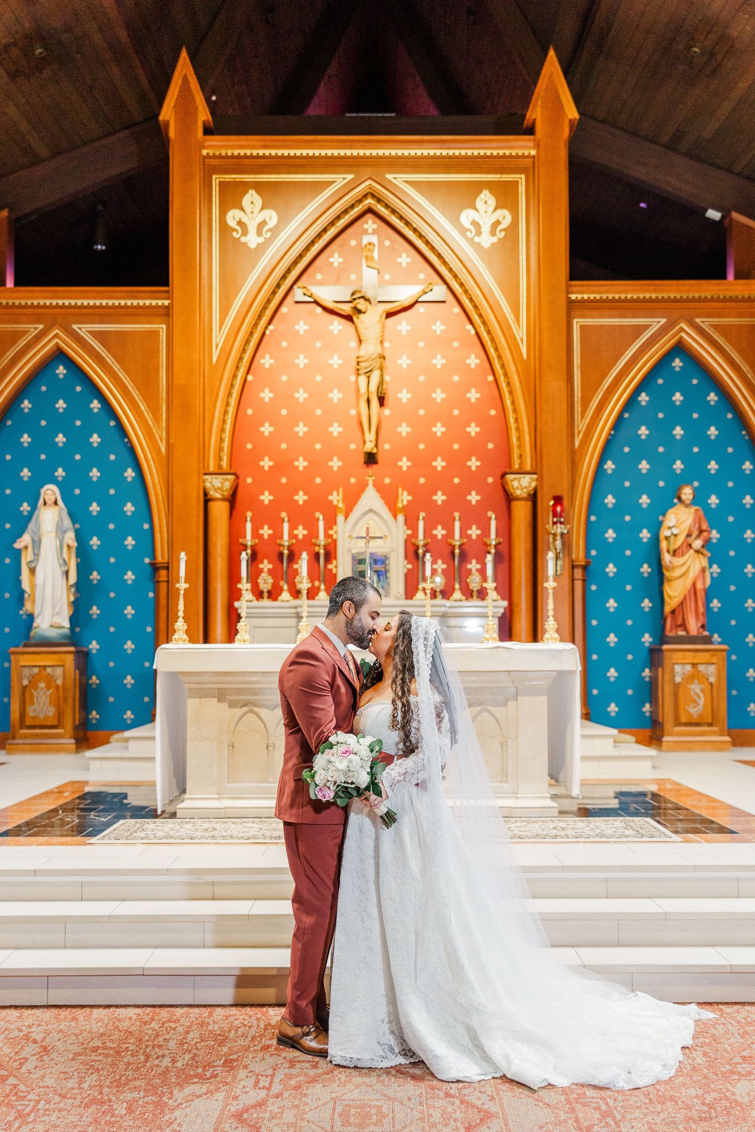 A wedding ceremony takes place in a Catholic church with ornate Gothic arches, blue and gold decor, and religious iconography.