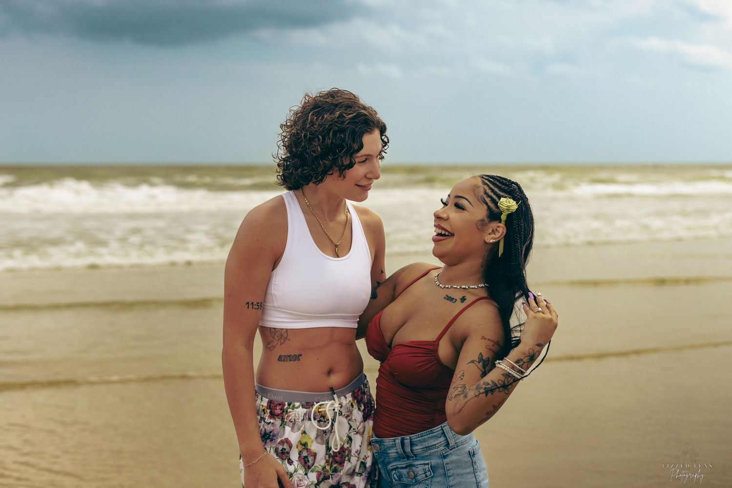 Two smiling women with tattoos share a joyful moment together on a sandy beach with ocean waves behind them.