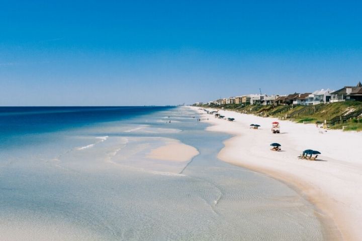 Aerial panoramic view of pristine white sand beach with turquoise waters and beachfront homes along the coastline.