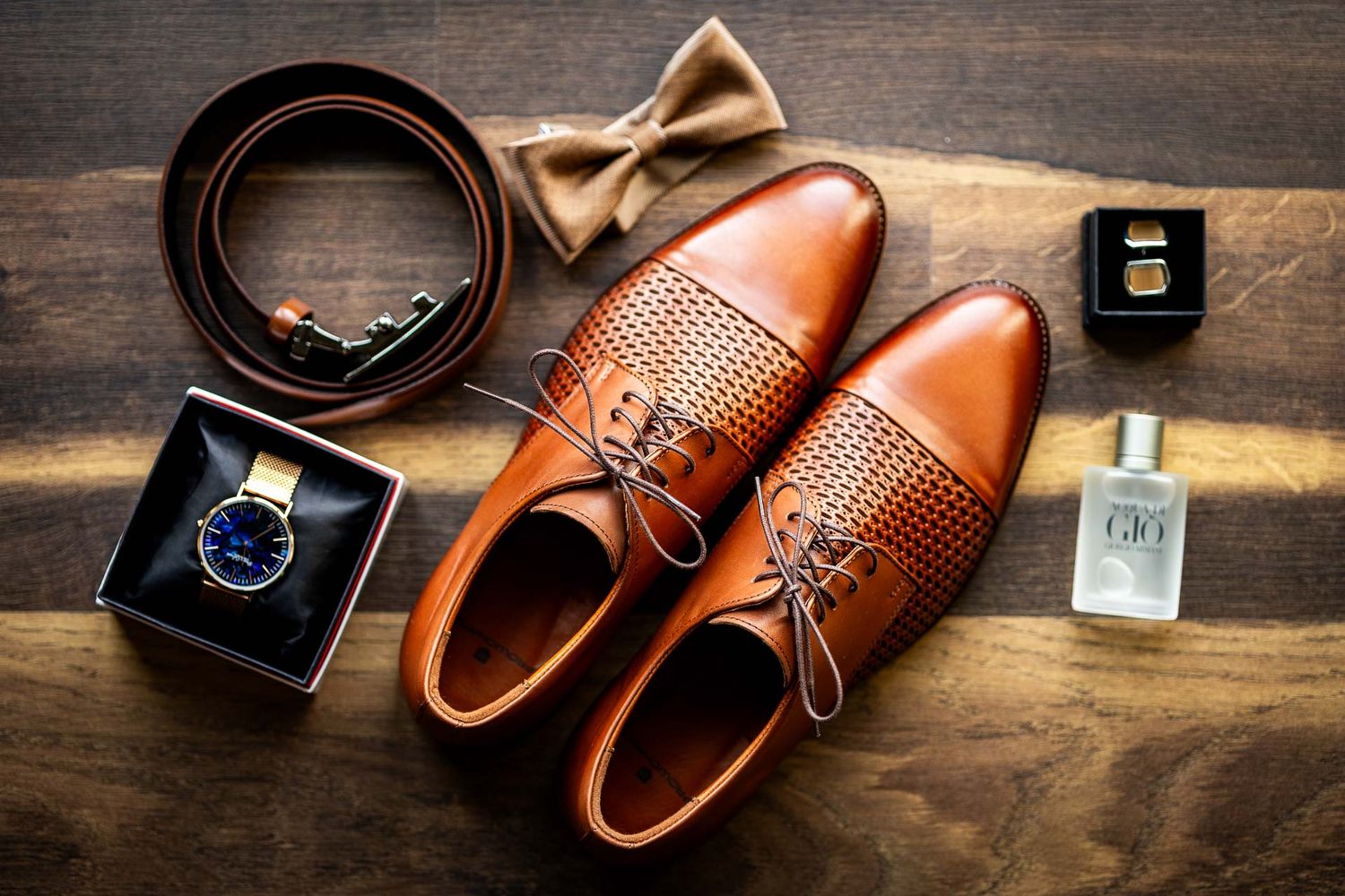 Brown leather dress shoes and accessories arranged on wooden surface for wedding photography.