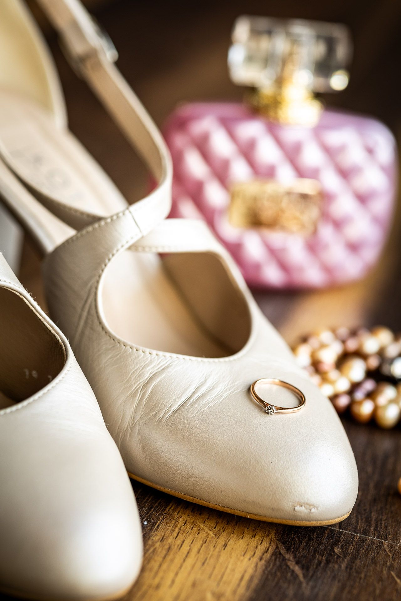 Close-up of white ballet flats with wedding ring and pink accessories on wooden surface.