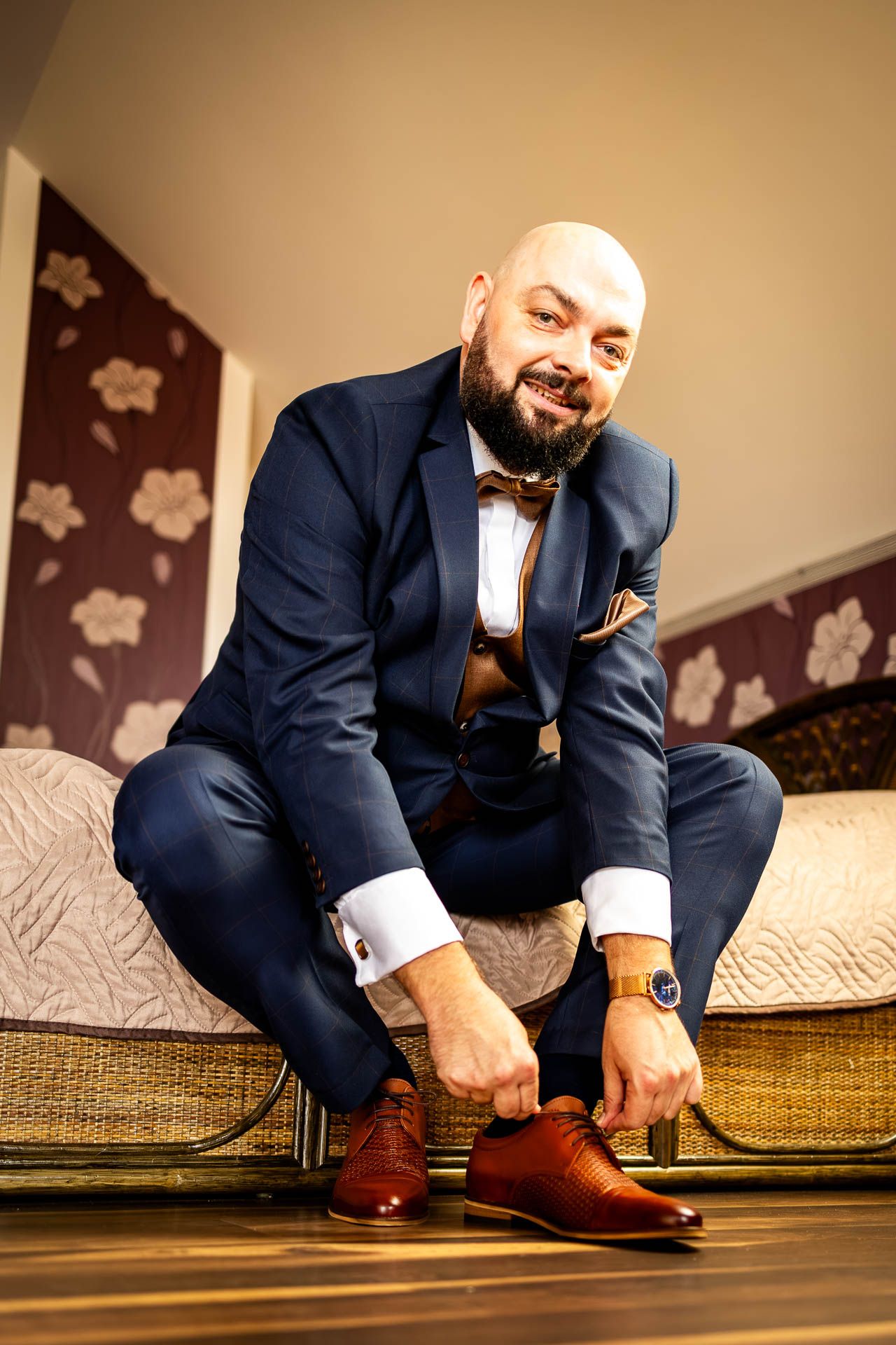 Groom in navy suit tying brown leather dress shoes while sitting on a couch.