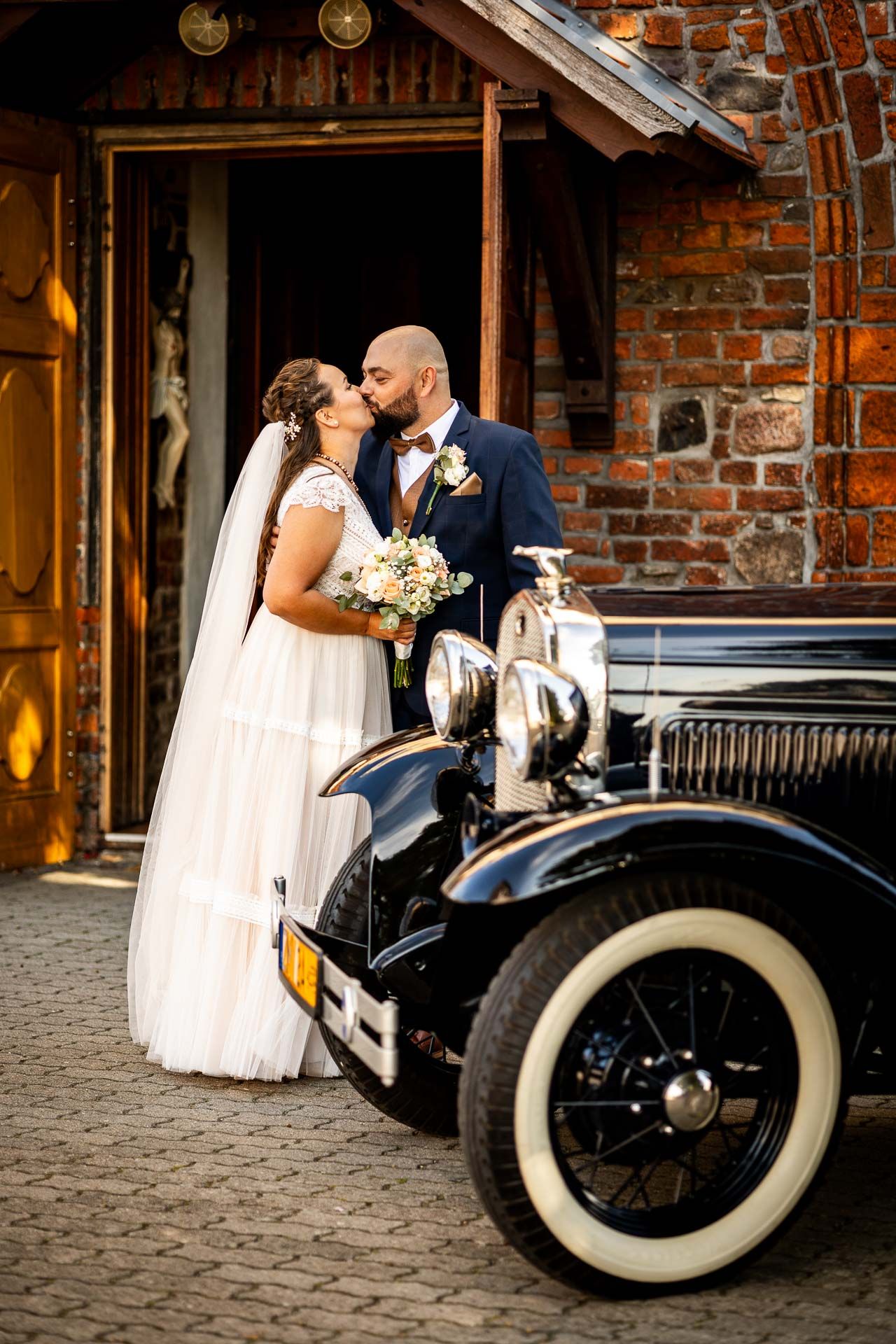 Wedding couple posing with classic vintage car against brick building.