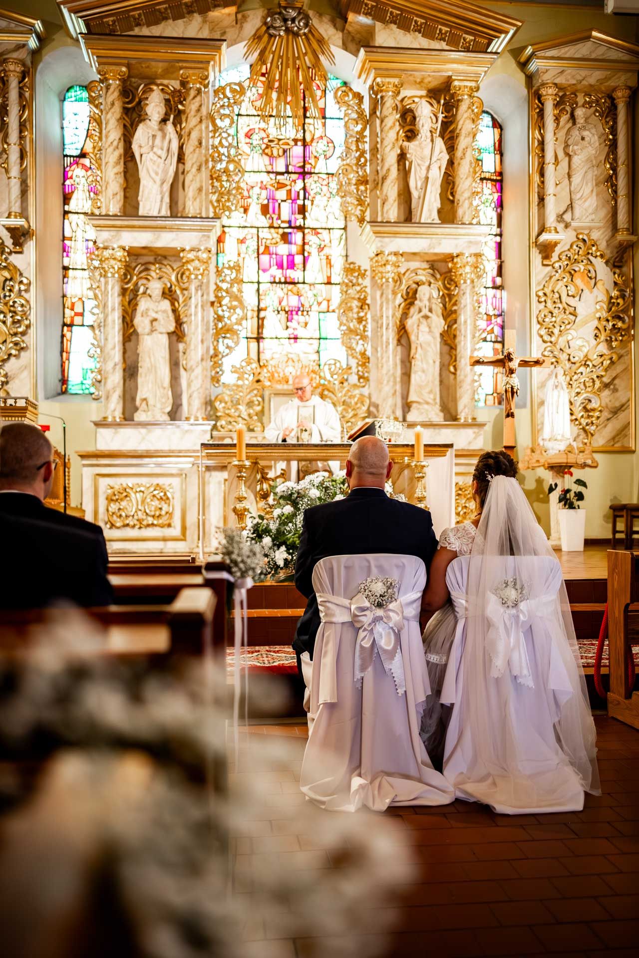 A wedding ceremony in an ornate church with stained glass windows and gilded statues.