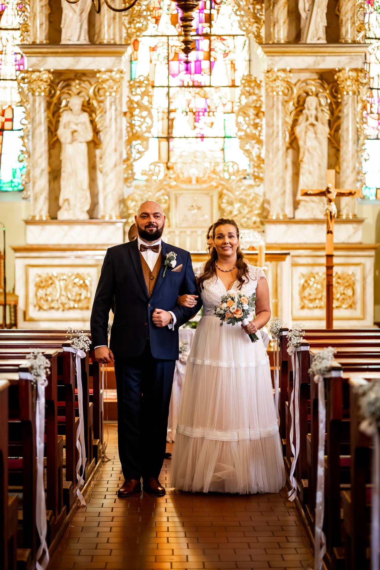 A wedding couple walks down the aisle of an ornate church with golden altar decorations.