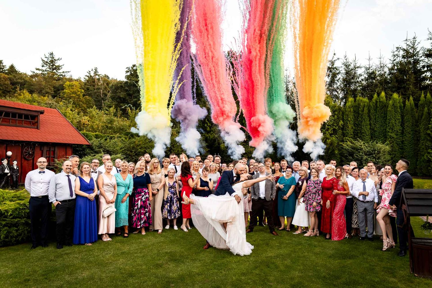 Wedding party and guests gathered outdoors for a group photo with colorful smoke bombs.