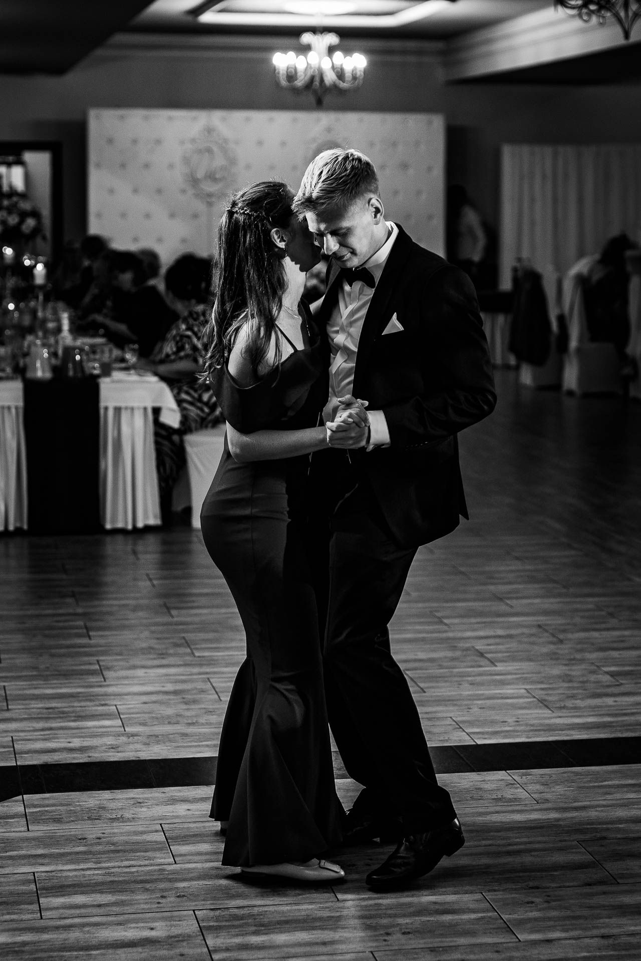 Black and white photo of couples dancing on a ballroom floor under chandelier lighting.