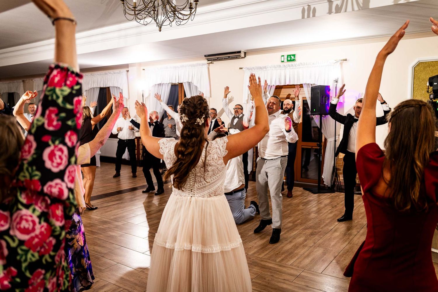Wedding guests dancing with raised arms on a dance floor under chandelier lighting at an indoor reception.