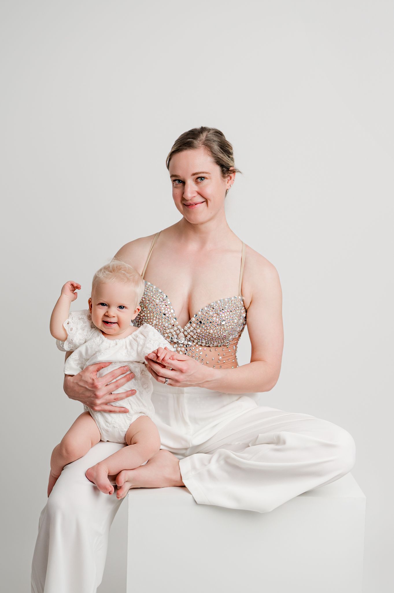 A parent in a sparkly top sits with a baby wearing white against a light gray studio backdrop.