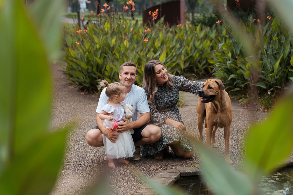 A family sits on a garden path with their brown dog while surrounded by tropical plants and greenery during a family photoshoot in sydney