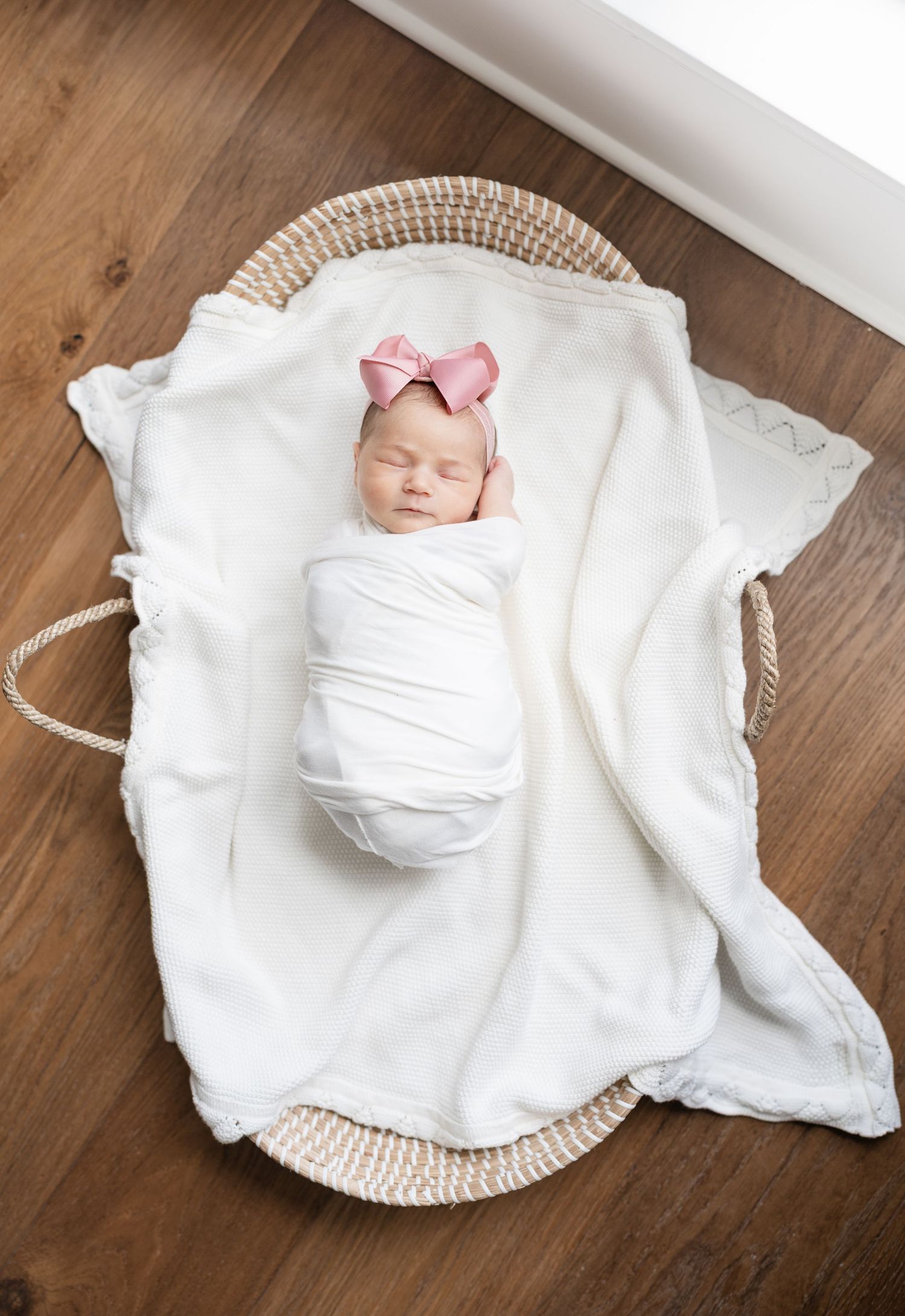 A sleeping newborn swaddled in white fabric rests on a round woven basket with a pink bow accent.