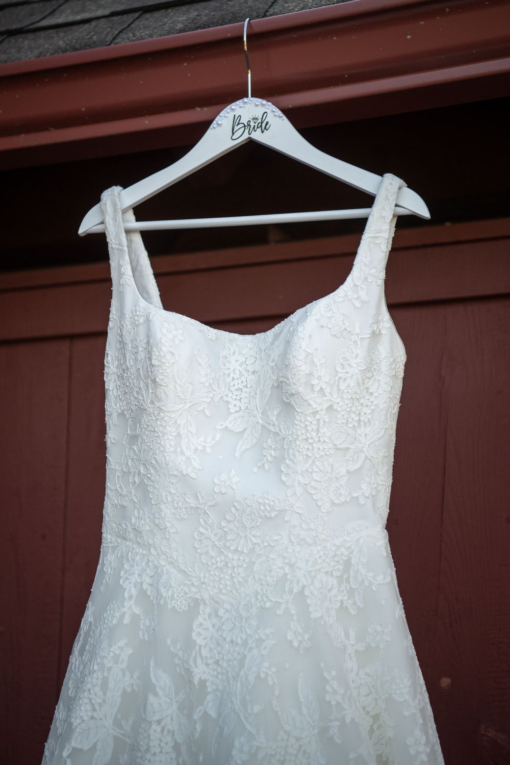 White wedding dress with lace details hanging on a personalized bride hanger against brown wall.