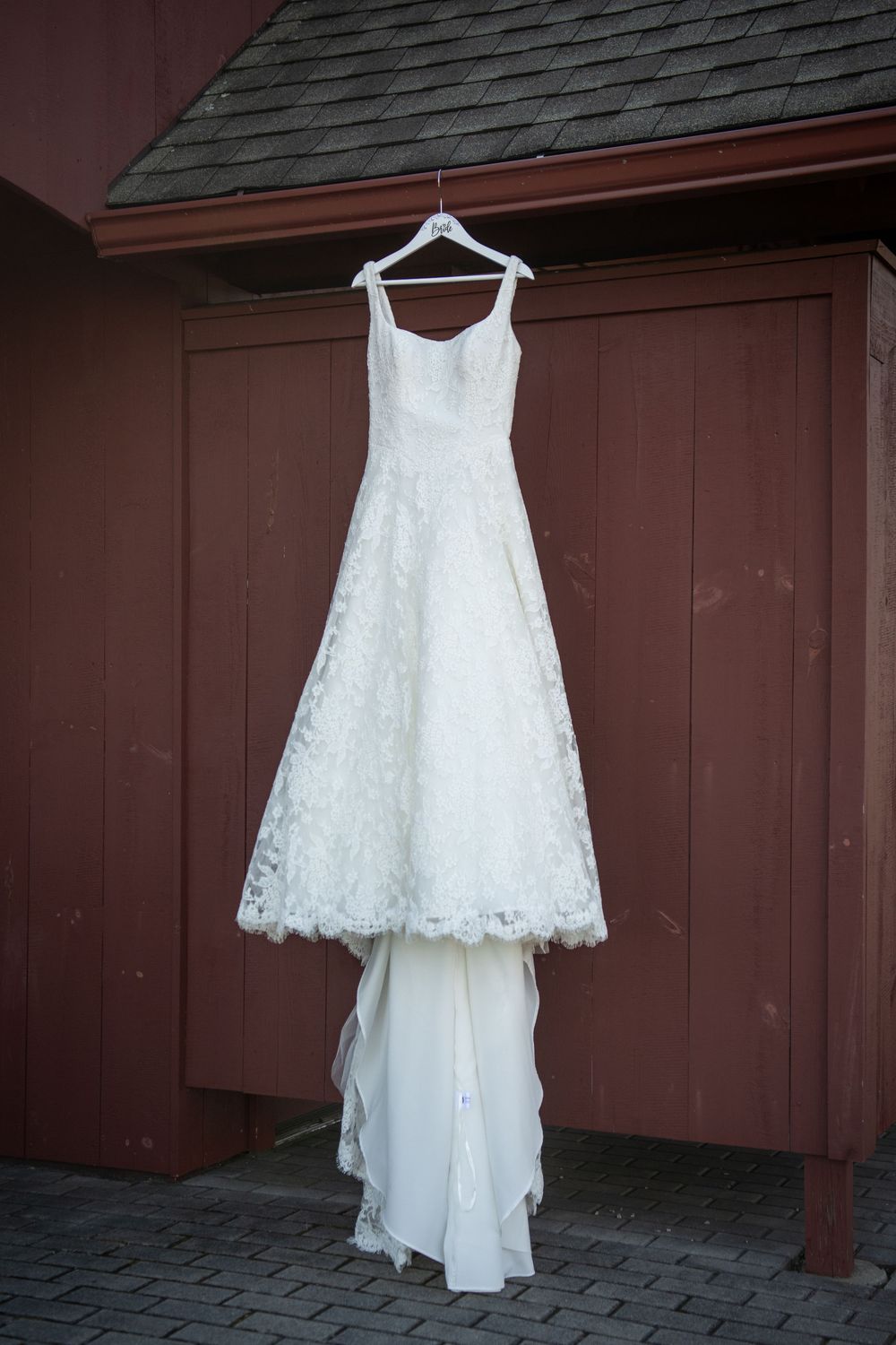 White lace wedding dress hanging against a dark brown wooden wall outdoors.