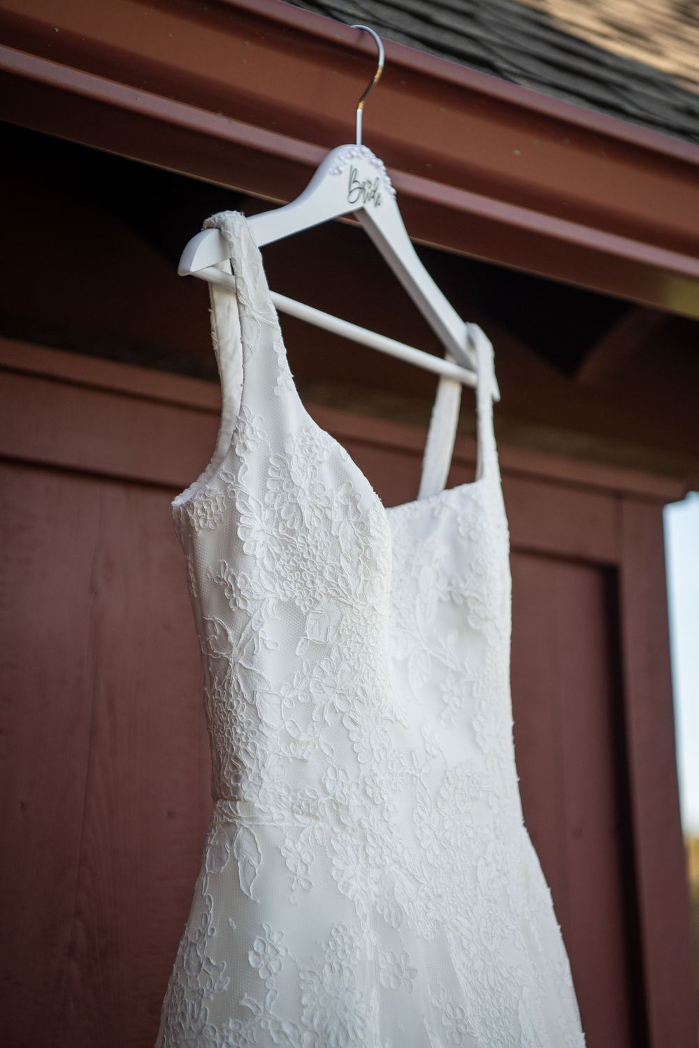 Elegant white wedding gown with intricate lace detailing displayed on a hanger against brown wall.