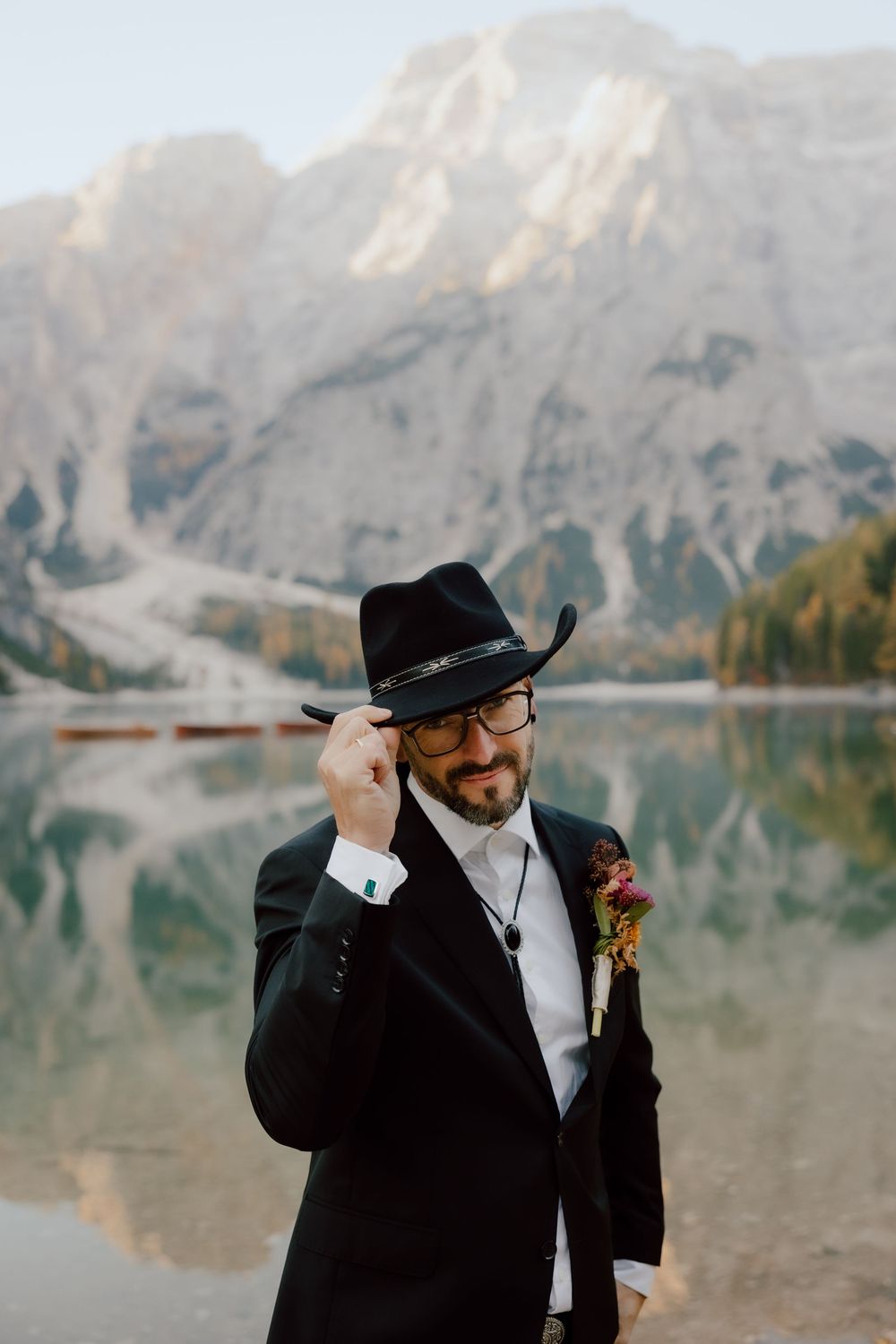 A groom in a black suit and cowboy hat poses against a mountain lake backdrop with dramatic peaks.