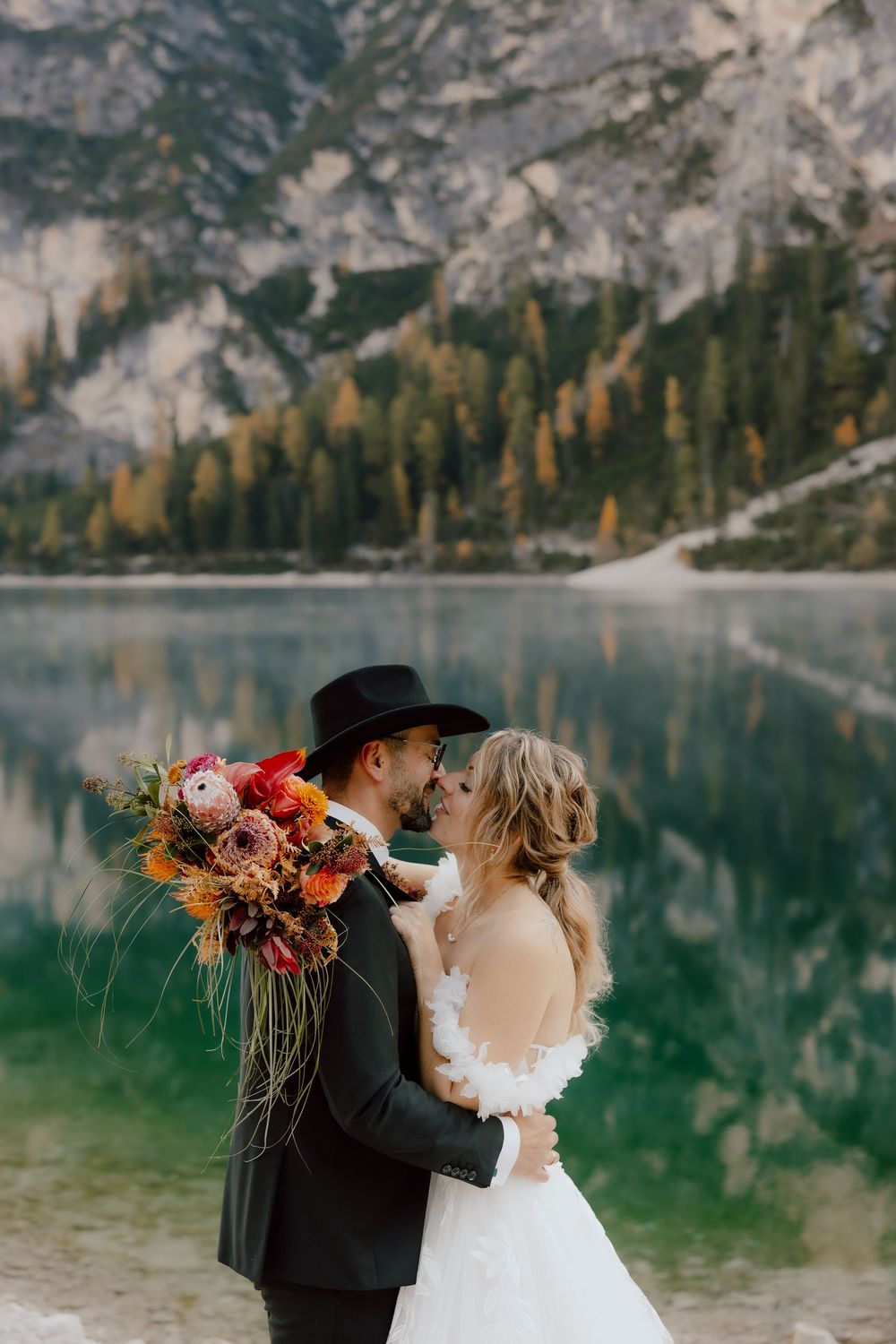 Scenic mountain lake wedding photo with colorful autumn bouquet against turquoise water and rocky cliffs.