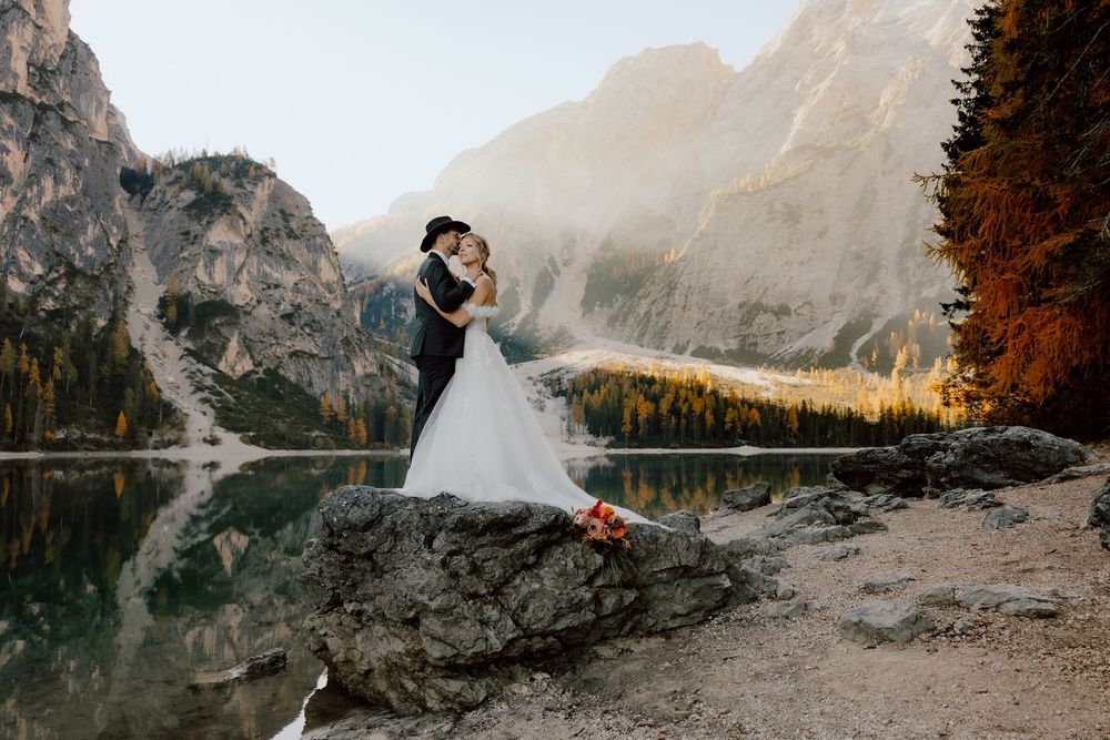 A couple in wedding attire poses along the rocky shores of a serene mountain lake at sunset.