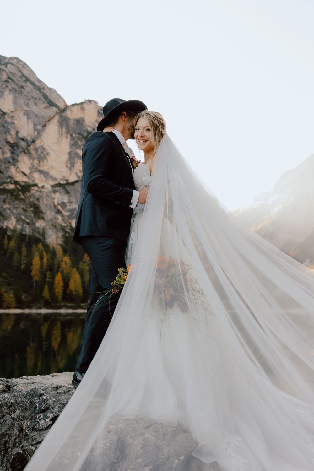 A wedding couple embraces on a rocky outcrop overlooking a mountain lake with a flowing white veil.