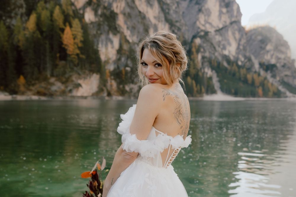 A bride in a white wedding dress poses against a backdrop of majestic mountain peaks and a serene alpine lake.